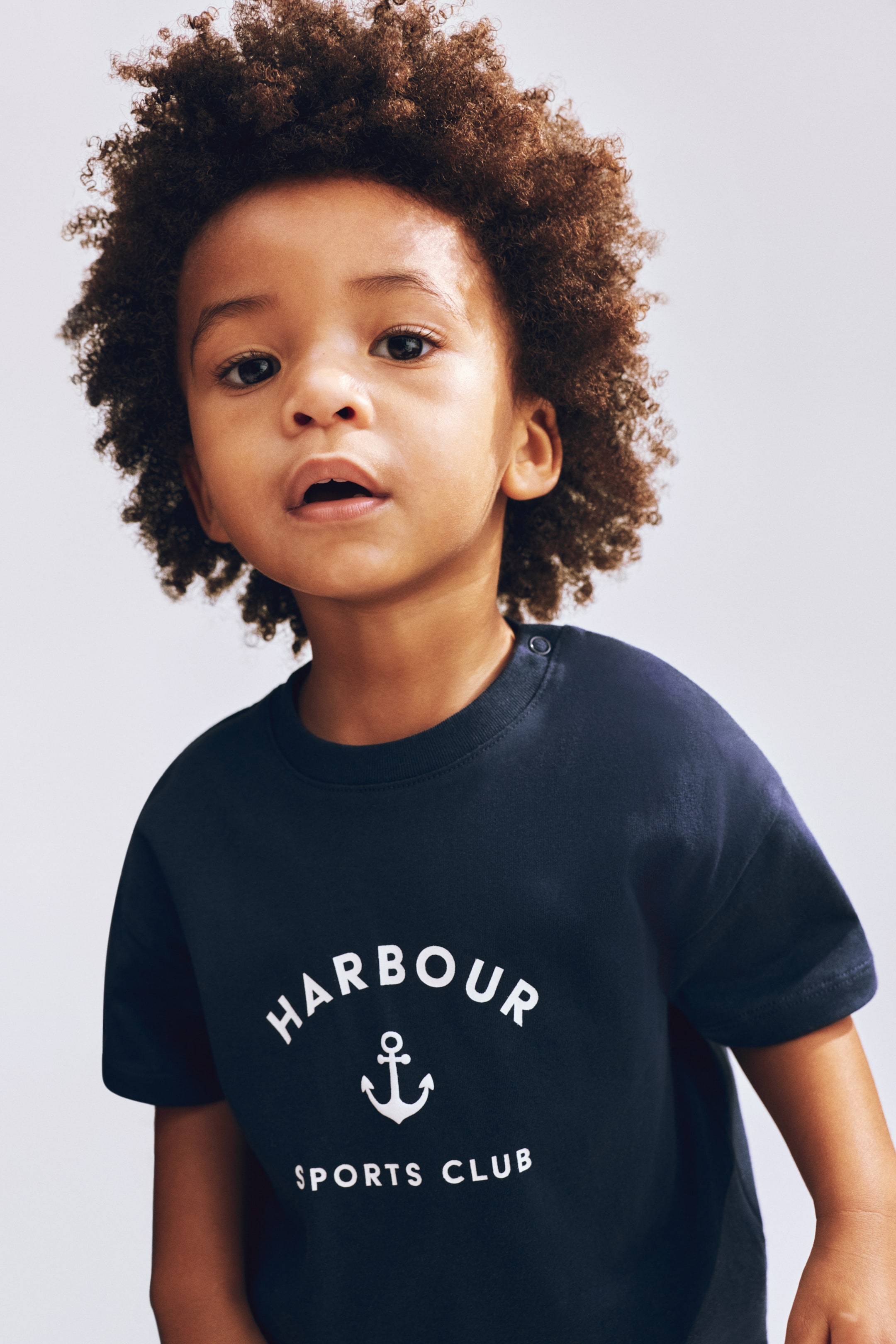View larger image: The dark blue t-shirt showcases a white graphic print on its front, featuring the word "HARBOUR" arched over an anchor symbol, with "SPORTS CLUB" positioned below. The t-shirt is crafted with a round neck and short sleeves, and two dark snap buttons secure the left shoulder.