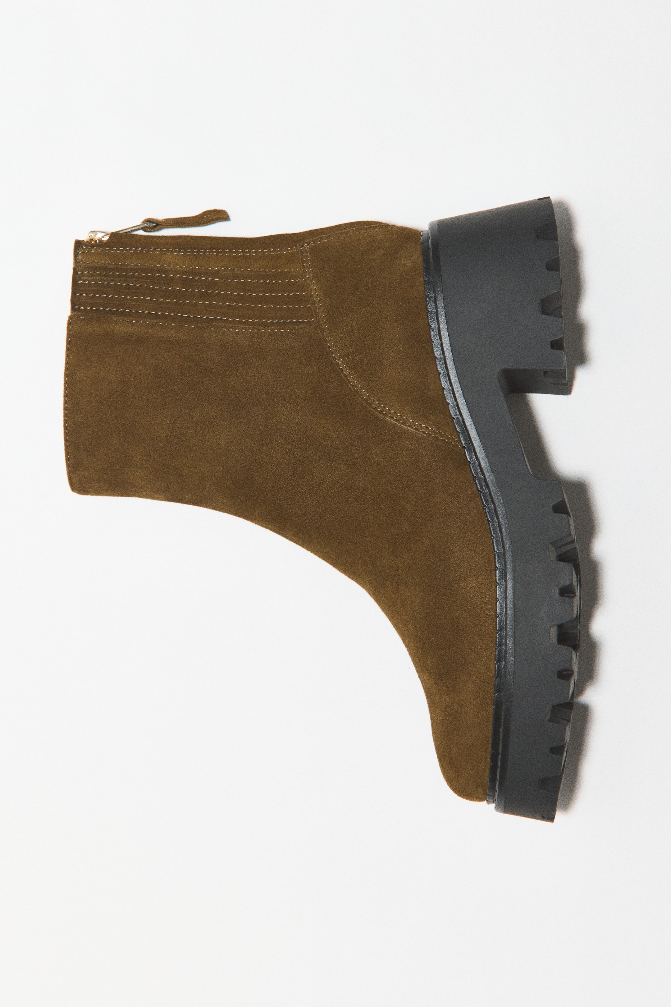View larger image: An olive green suede ankle boot is shown in side profile, featuring a rounded toe and a chunky black block heel with a patterned lug sole. The boot is designed with elasticated stitching at the side-back collar and a zipper at the back with a matching suede pull tab.