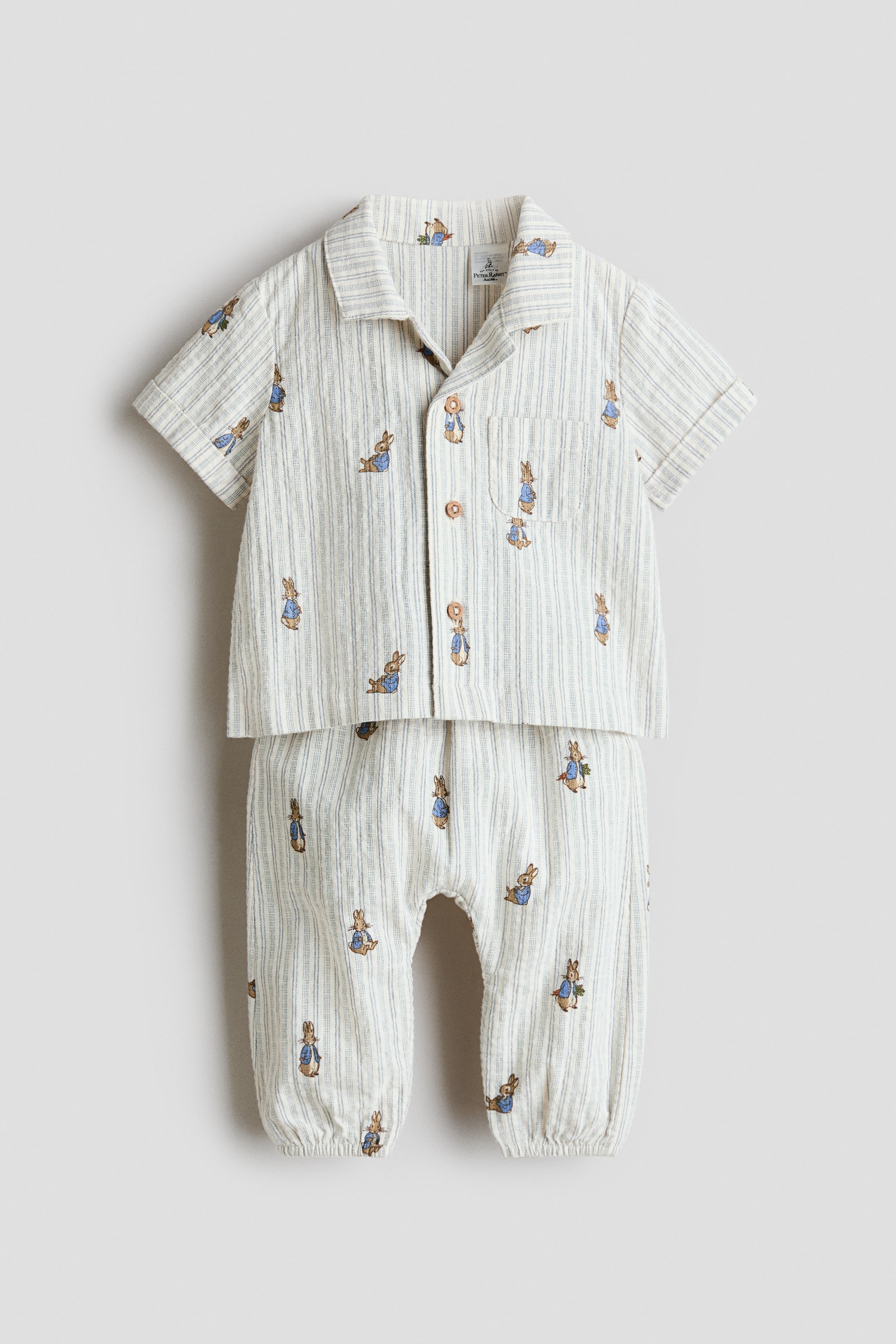 Agrandir l'image: Ce set, présenté de FACE, comprend une chemise et un pantalon, confectionnés dans un tissage de coton doux et texturé avec un motif rayé et un imprimé intégral de personnages de lapin. La chemise est conçue avec un col de style resort, des boutons sur le devant, une poche plaquée ouverte et des manches courtes avec des revers cousus aux poignets, tandis que le pantalon présente une élastication recouverte à la taille et aux ourlets.