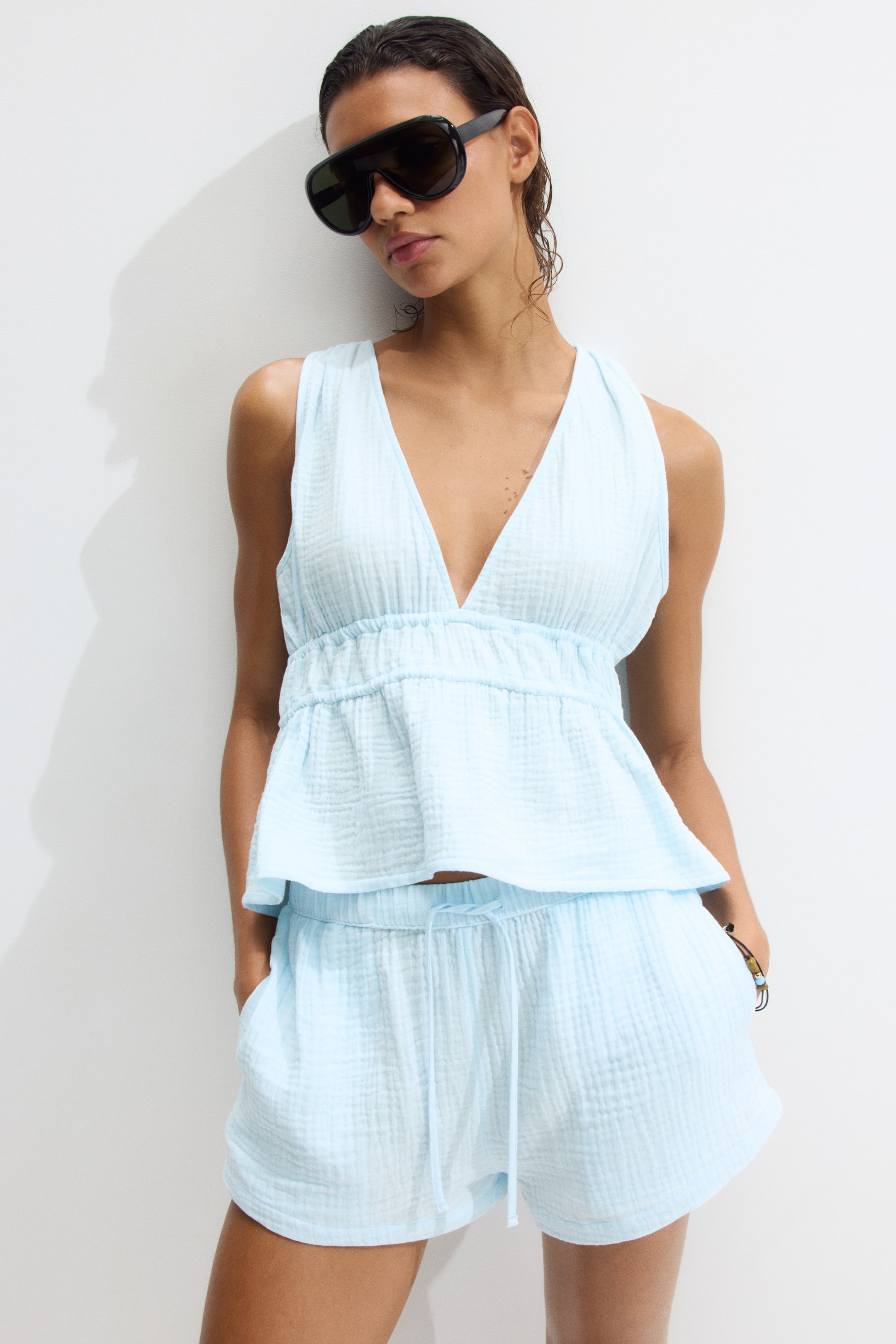View larger image: Loose-fit beach shorts in airy light blue cotton muslin are being worn by the model. They are featuring an elasticated, drawstring waist and discreet side pockets, and the model is placing one hand in a pocket.