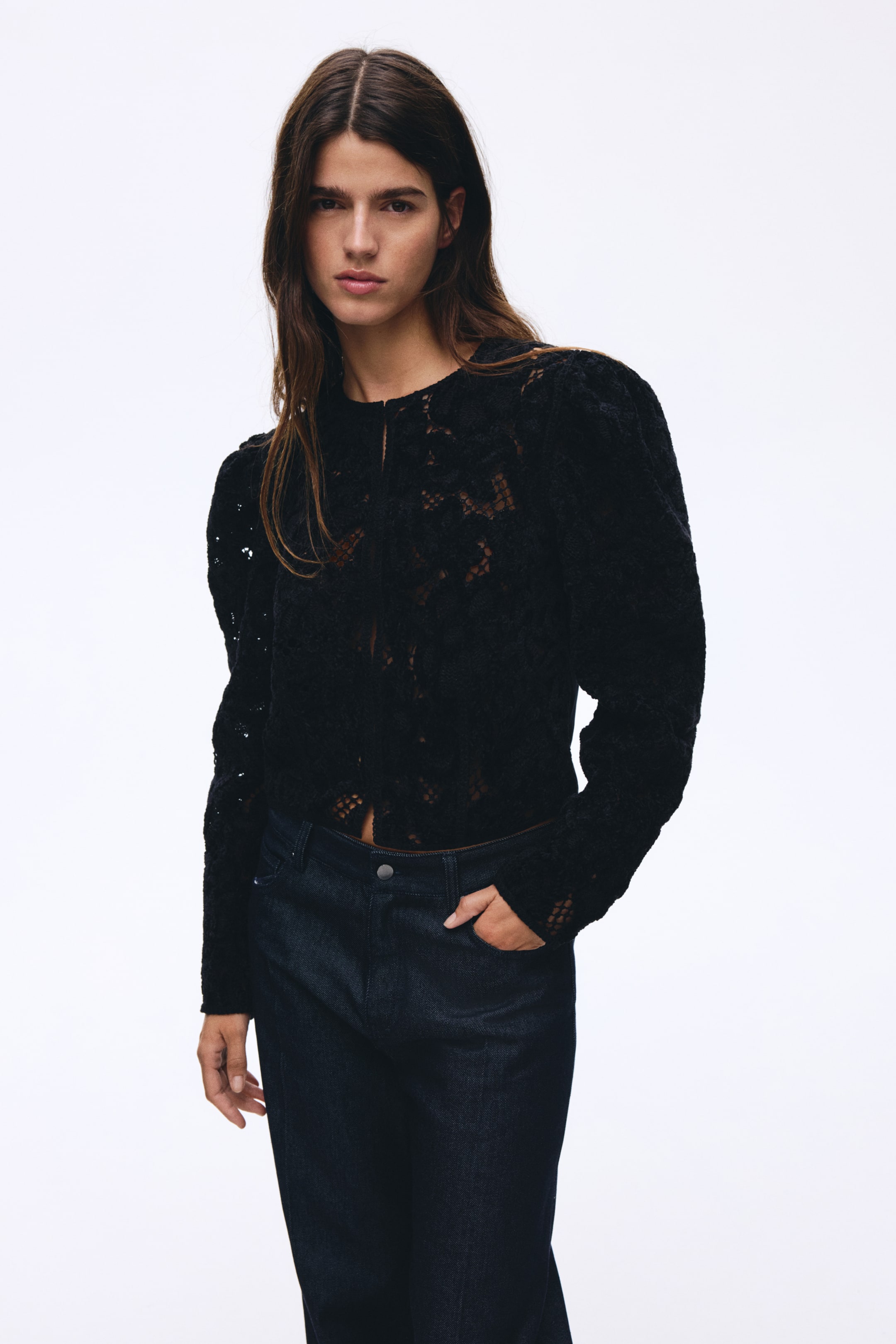 Größeres Bild ansehen: Ein schwarzes Spitzentop wird von vorne präsentiert, mit einem runden Halsausschnitt, langen Ärmeln mit Puffschultern und einem aufwendigen Lochstrickmuster, das knapp oberhalb des Bunds von dunklen Jeans sitzt.