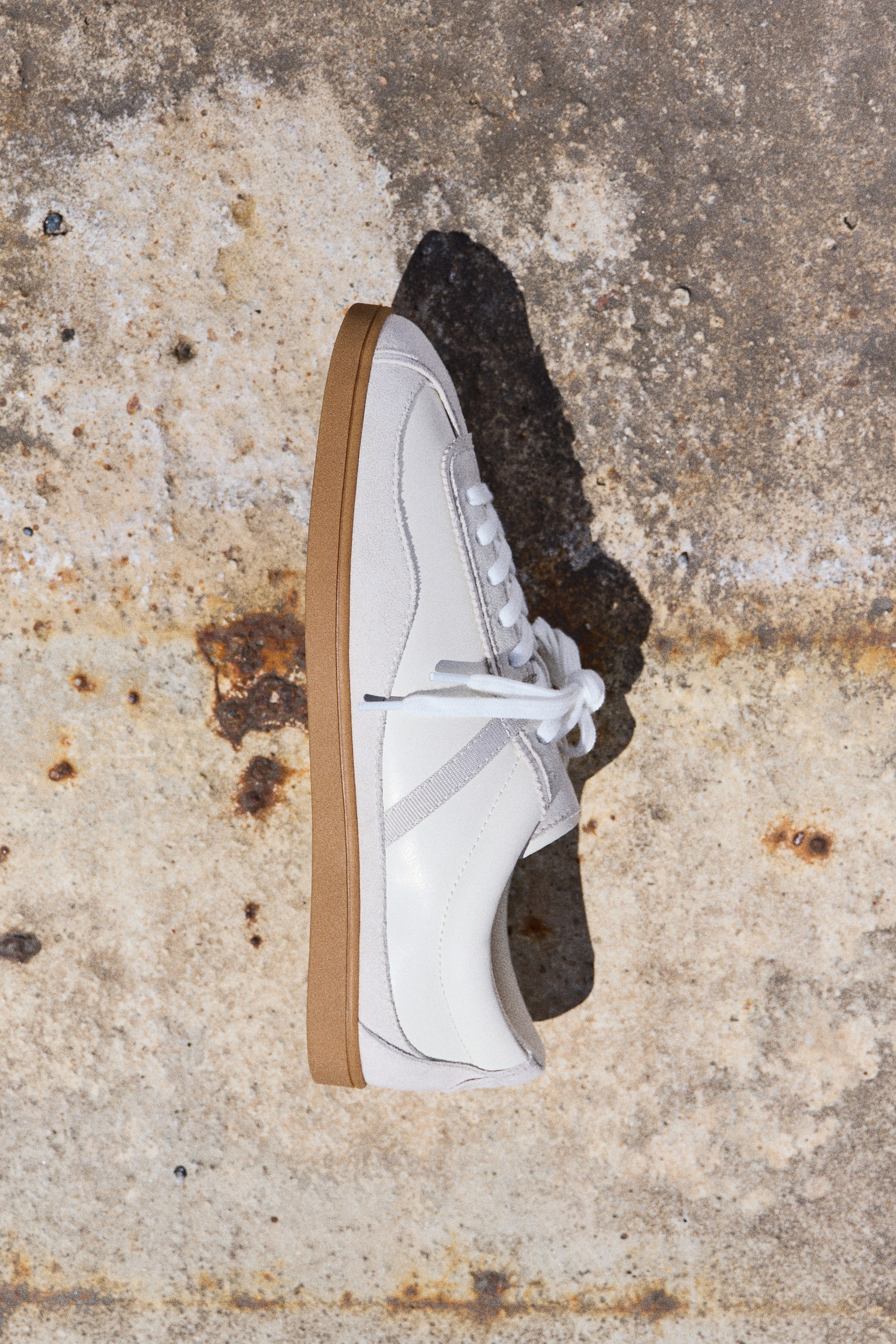 Vis større bilde: Lave sneakers viser sin ytre side, med en lysfarget belagt stoffoverdel som har myke, børstede detaljer og en subtil teksturert side stripe. Hvite lisser er synlige foran, og en kontrastfarget brun gummisåle fullfører designet.