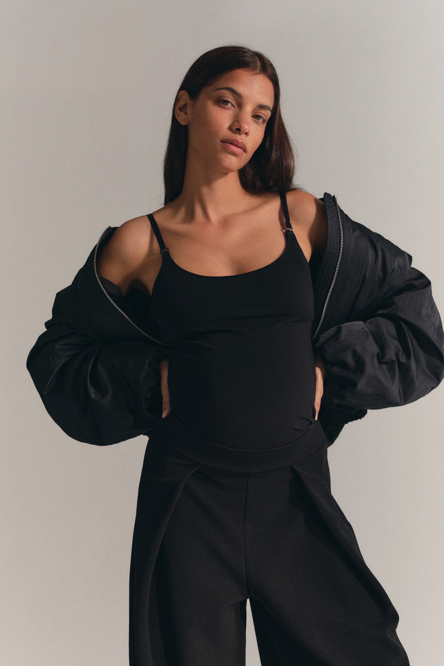 A black nursing cami with a scoop neck and narrow, adjustable straps with visible nursing clasps is worn by the model. The cami is layered under an open black jacket worn off the shoulders and paired with black trousers. - 1