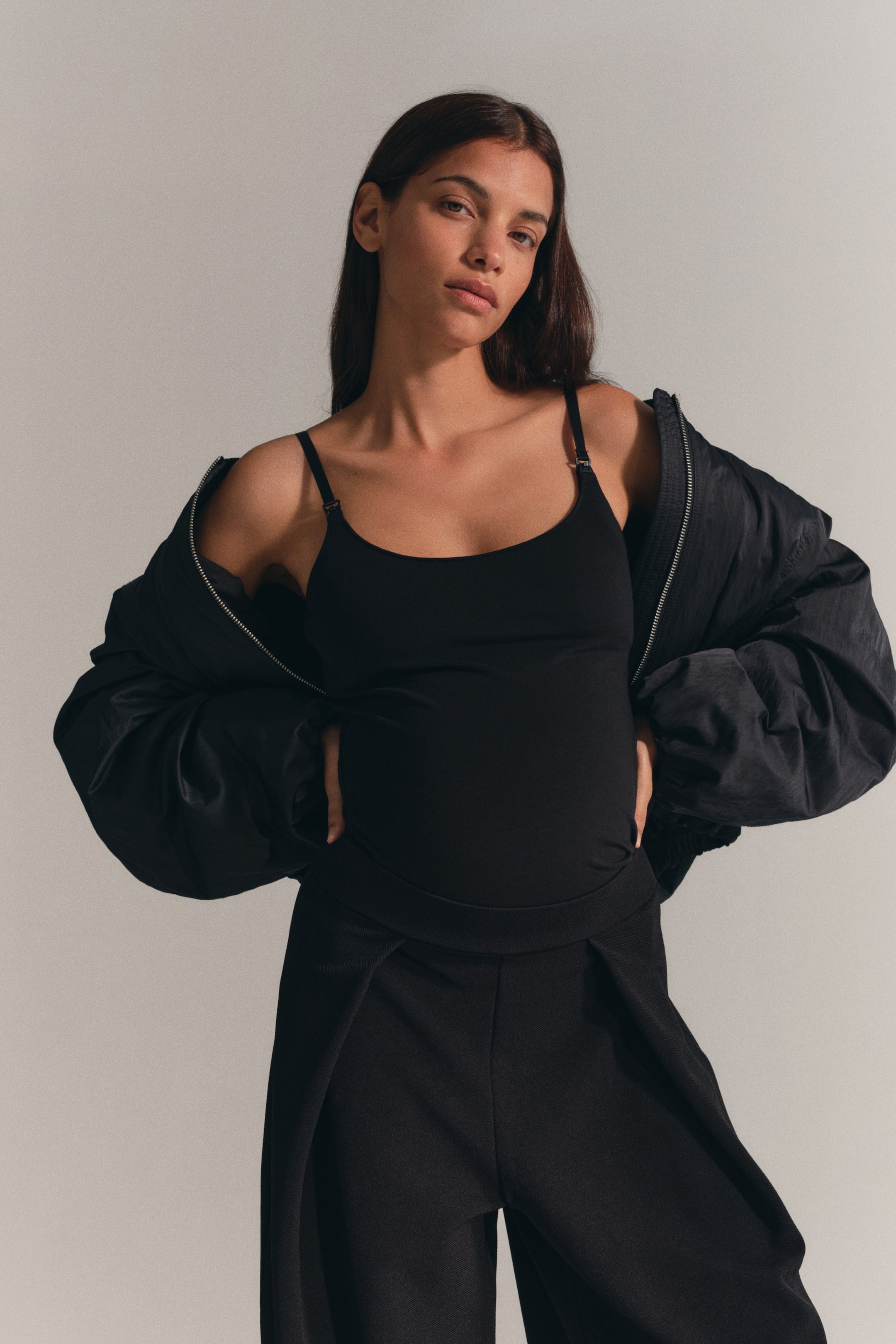 Ver imagem maior: Uma camisola de alças de amamentação preta, com decote redondo e alças finas ajustáveis com fechos de amamentação visíveis, está a ser usada pela modelo. A camisola está sobreposta sob um casaco preto aberto, usado caído sobre os ombros, e combinada com calças pretas.