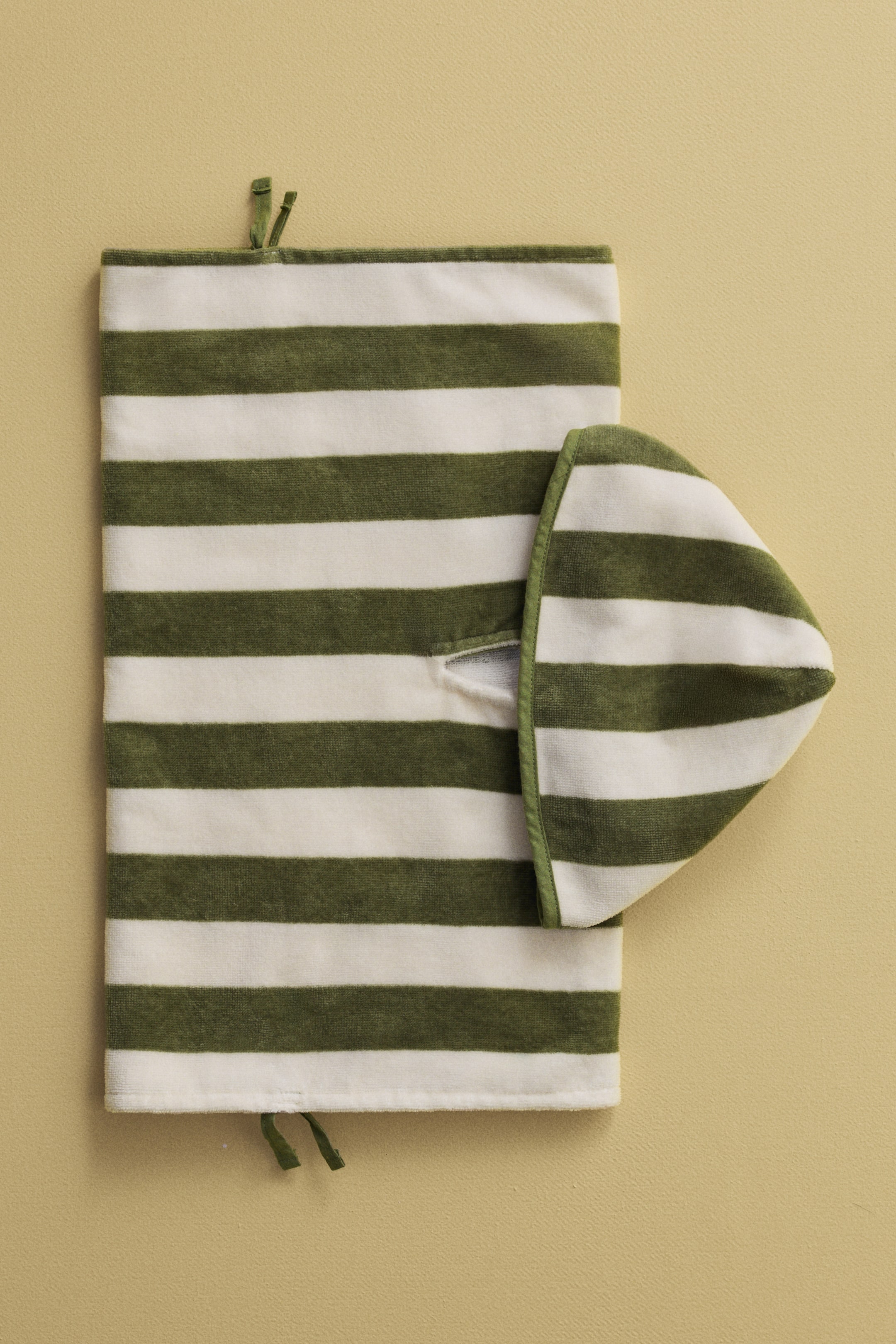 Nagyobb kép megtekintése: Egy poncsó törölköző, amely az elejét mutatja, laposan fekszik, és váltakozó plüss olíva zöld és törtfehér vízszintes csíkokkal rendelkezik. Egy hozzáillő csíkos kapucni van a jobb felső oldalra hajtva, a bal oldali szél tetején és alján keskeny olíva zöld kötők láthatók.