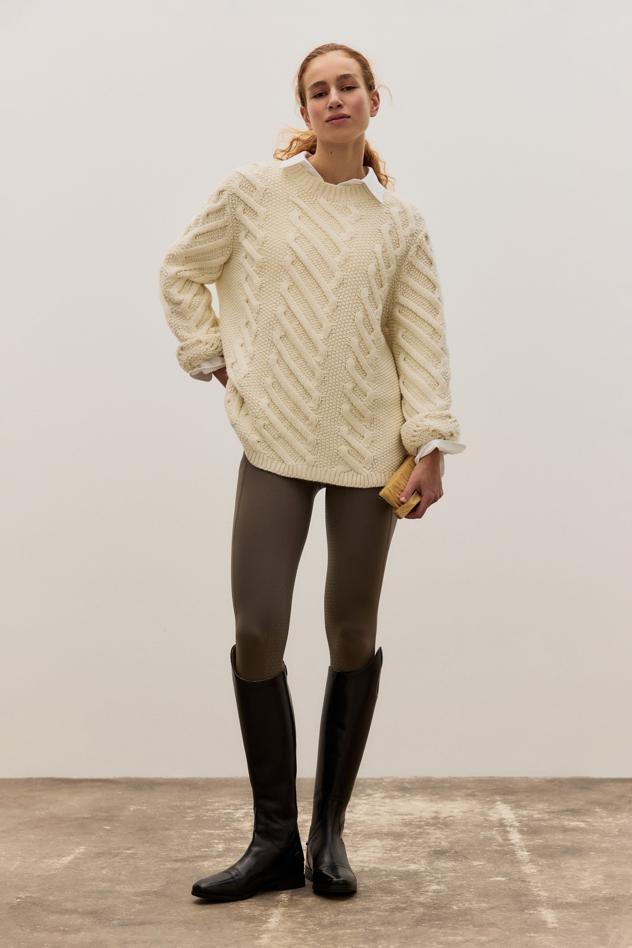 Zobrazit větší obrázek: Krémový svetr s copánkovým vzorem představuje velké diagonální copánkové vzory na přední straně a rukávech, s žebrovanými manžetami. Pod svetrem je viditelně vrstven límec a manžety bílé košile.