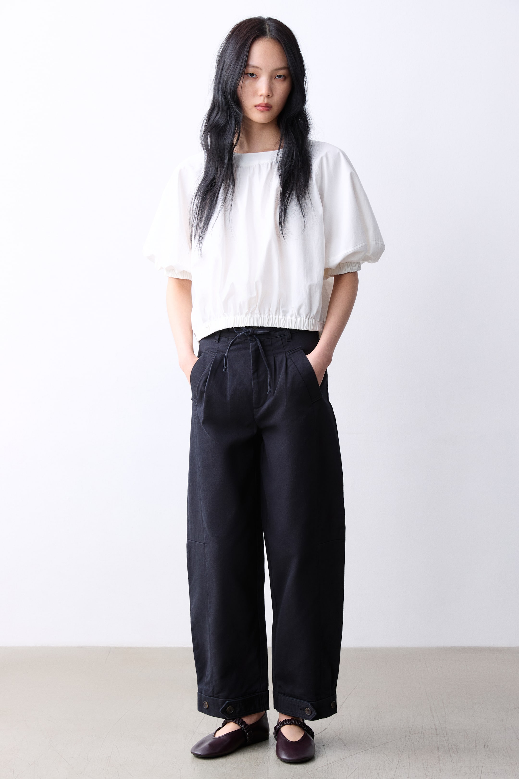 拡大画像を表示: 白いクロップドトップは、ギャザー入りのボートネックライン、パフショートスリーブ、そしてゴム入りの裾が特徴です。スタイリングには、ドローストリングウエスト、フロントプリーツ、ボタン付きカフスが施された黒のワイドレッグトラウザーズと、ギャザーストラップが付いたダークマルーンのバレエフラットが組み合わされています。