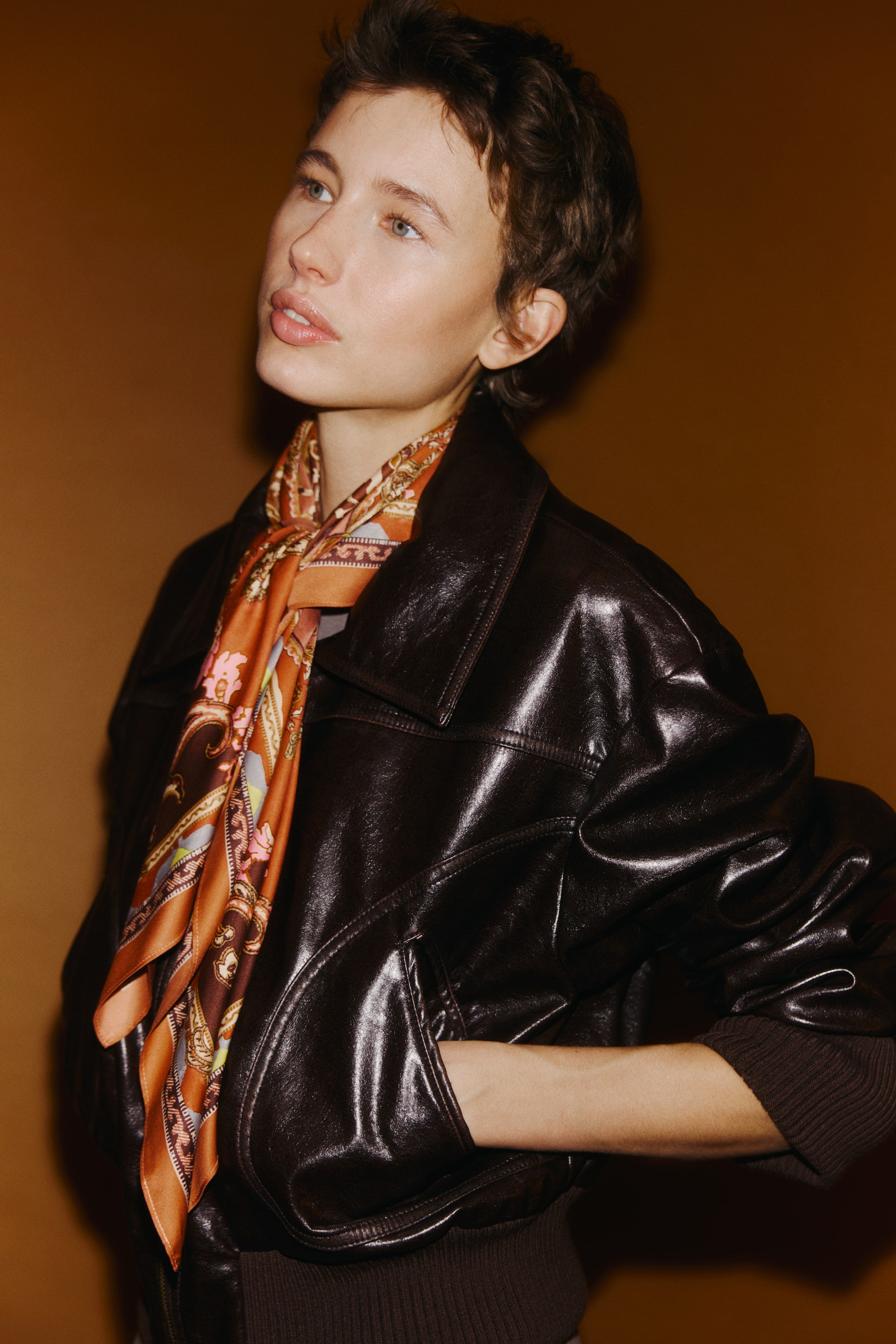 View larger image: A dark brown, shiny bomber jacket features a pointed collar, front welt pockets, ribbed cuffs, and a ribbed waistband. A multi-patterned silk scarf, predominantly orange and brown, is tied around the jacket's collar.