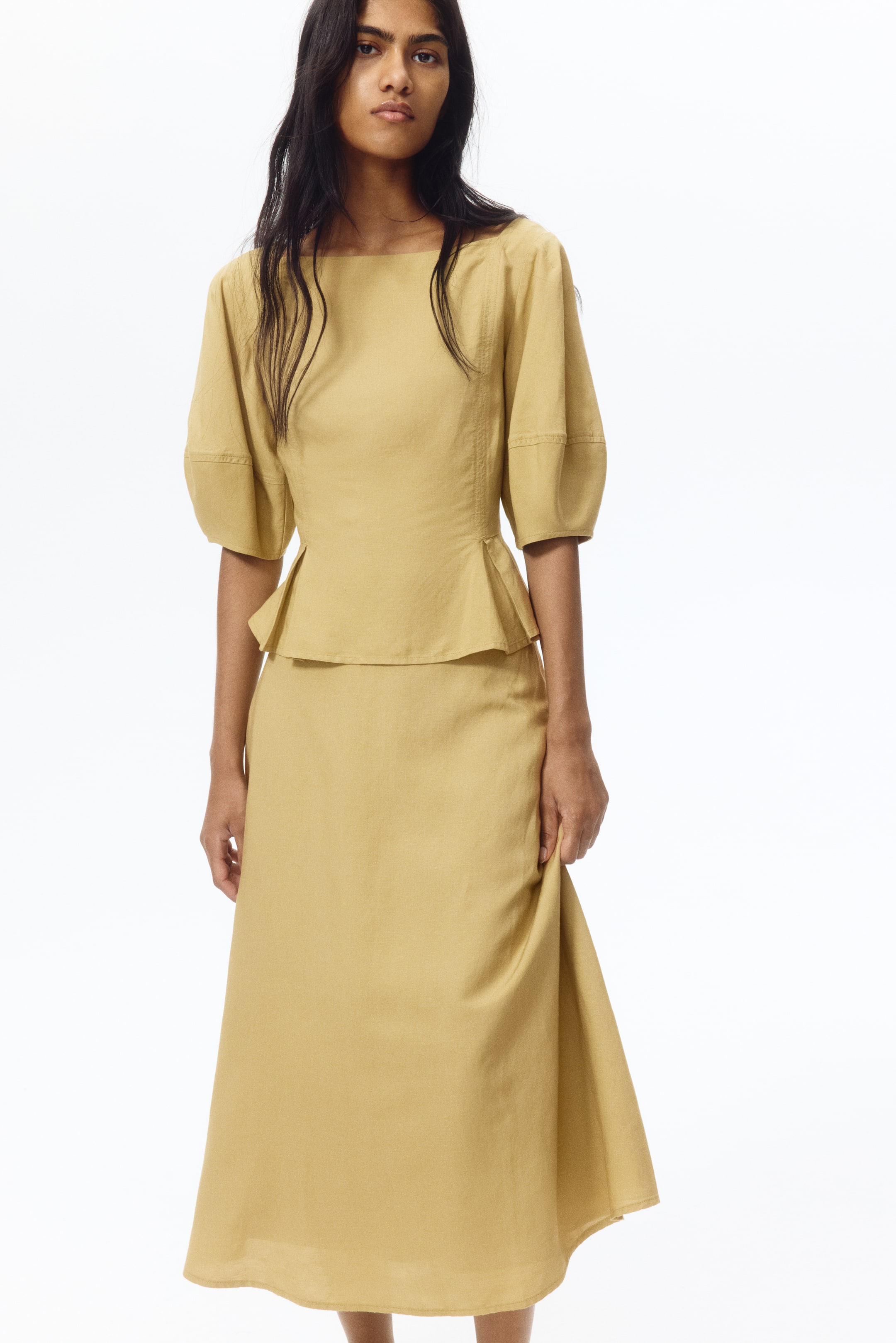 View larger image: Presenting a mustard yellow two-piece ensemble, comprising a peplum top and a coordinated midi skirt. The top showcases a square neckline, elbow-length puffed sleeves with banded cuffs, and a fitted bodice with a flared peplum hem. The flowing midi skirt is elegantly displayed to highlight its graceful drape.