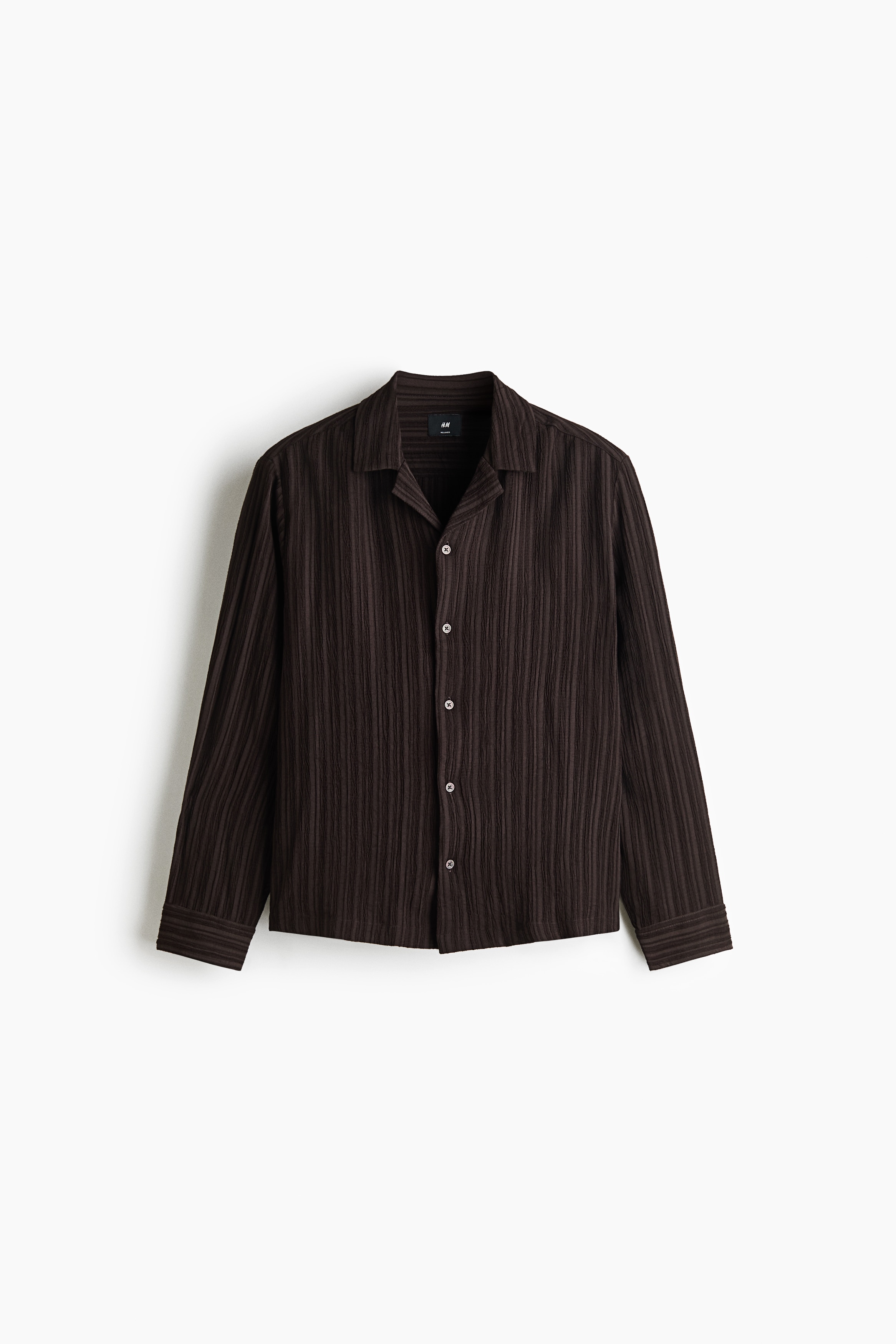 A dark brown, vertically textured long-sleeve shirt in a relaxed fit is worn by the model. The shirt features a resort collar and is styled with a partial tuck into dark trousers secured with a belt.