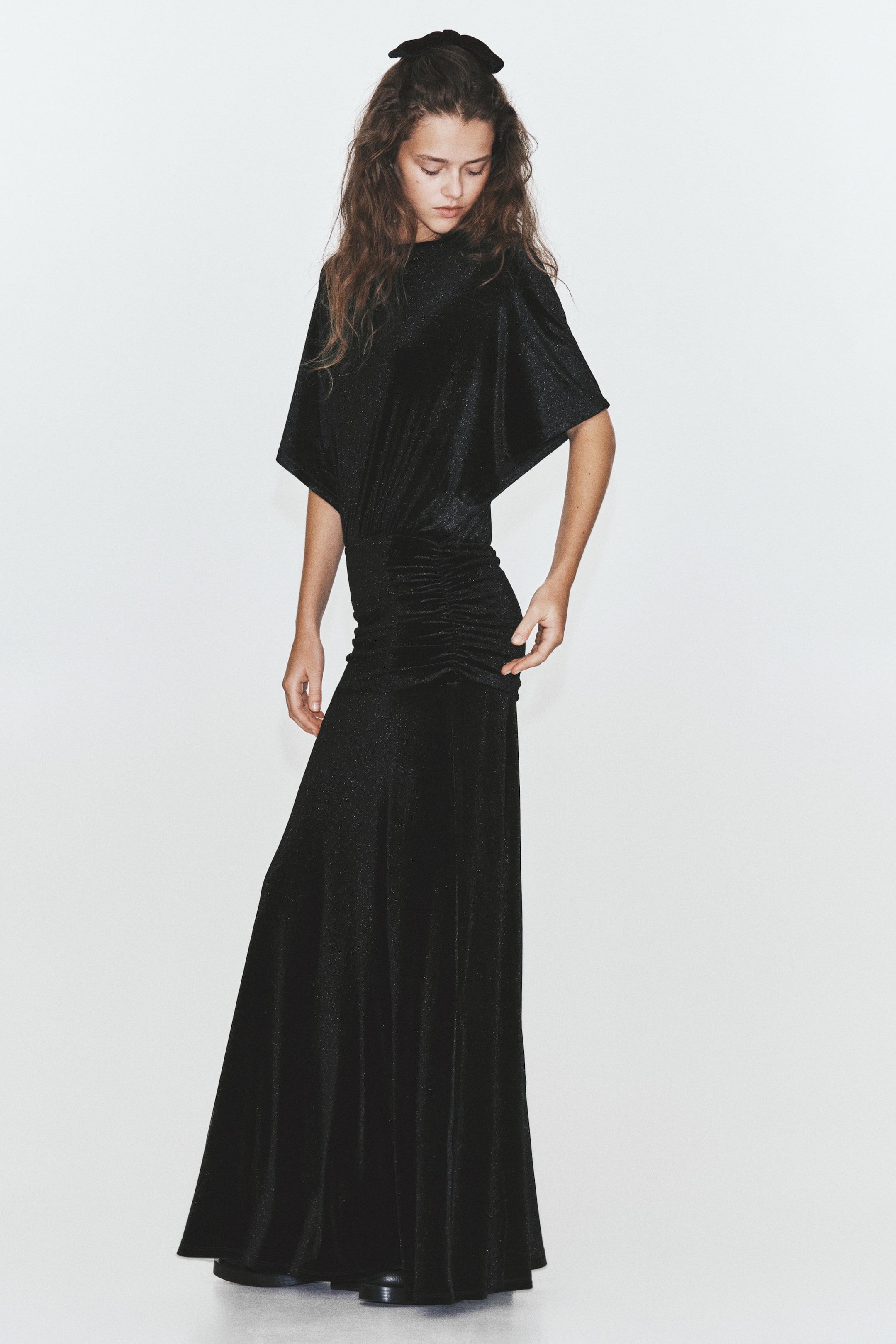 View larger image: A black velvet maxi dress featuring a high crew neckline, draped butterfly sleeves, and a gathered, ruched waist is being worn by the model. The model is standing while lightly touching the side of the ruched waistline.