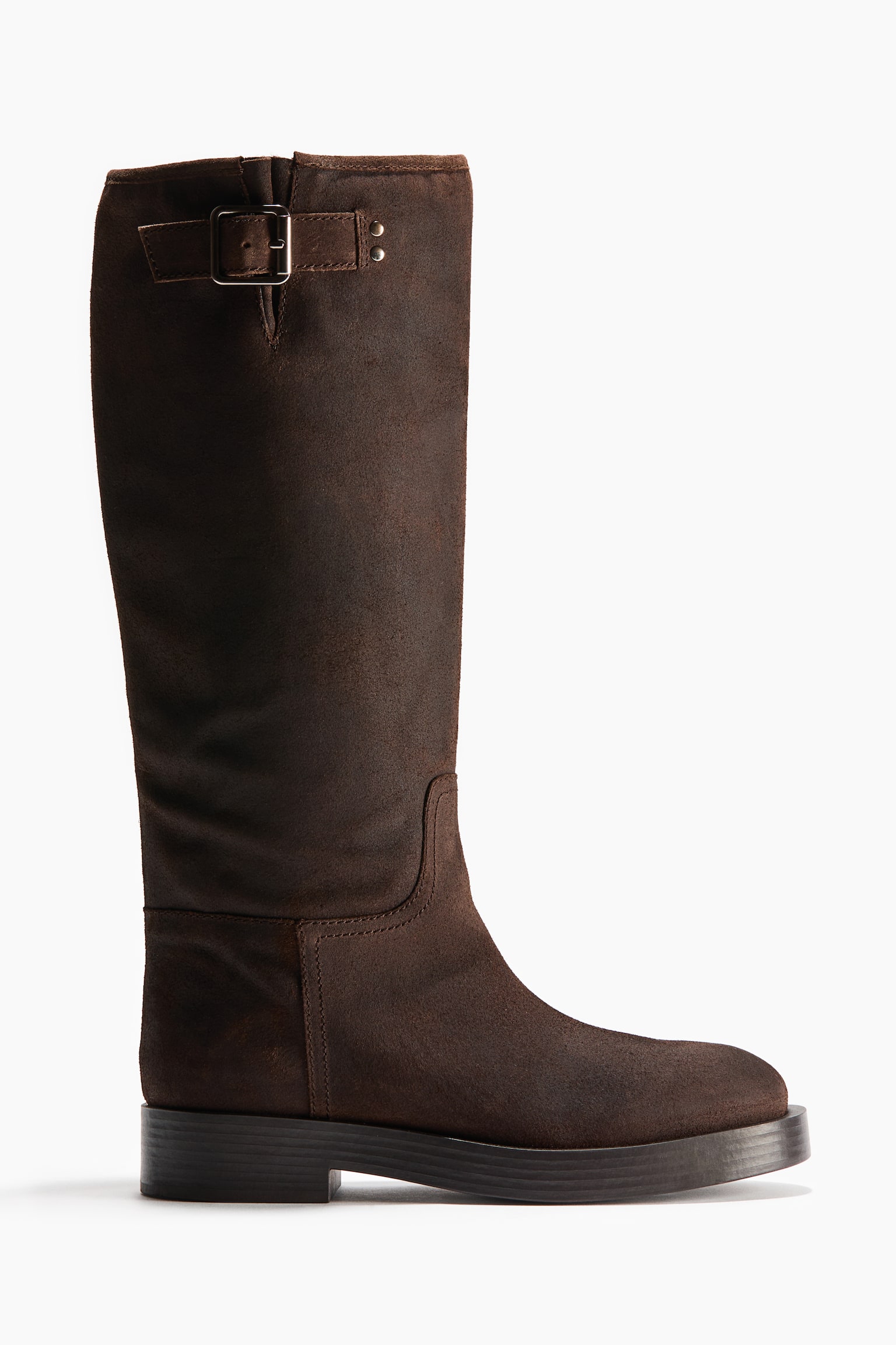A dark brown suede knee-high boot is shown in side profile facing right, featuring a rounded toe and a thick, flat black sole. A decorative strap with a silver-toned metal buckle and small studs details the upper collar of the wide shaft.