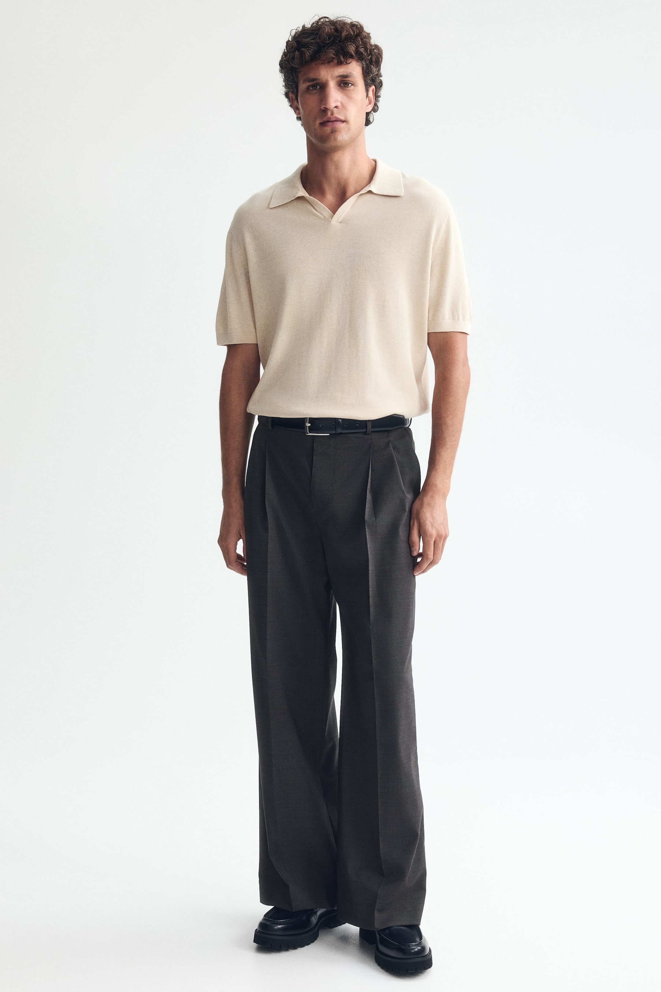 View larger image: The model is wearing tailored dark gray trousers, featuring pleats at the top, creases down the legs, and a loose fit. A black belt secures the trousers at the waist.