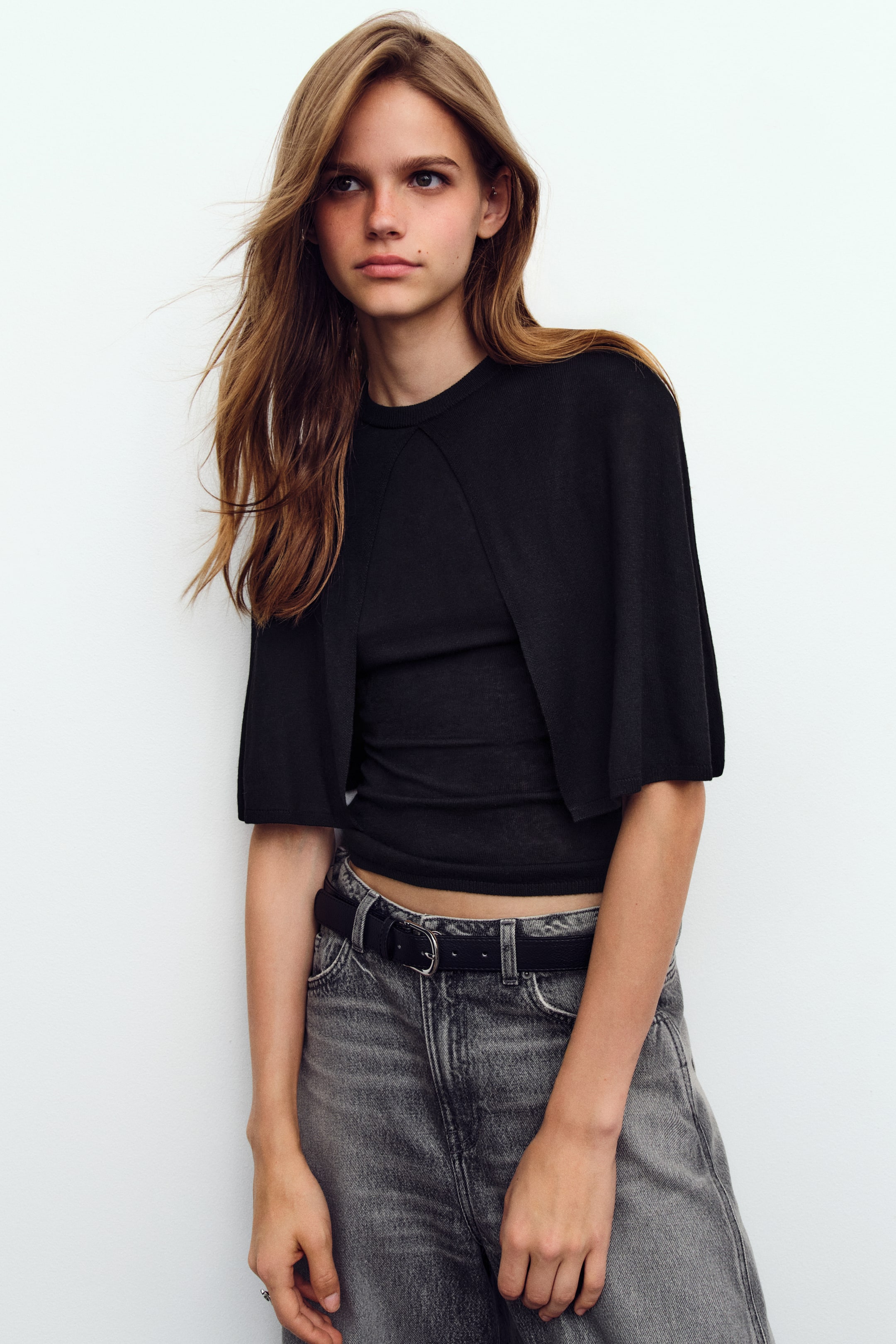 Ampliar la imagen: La modelo lleva una blusa negra ajustada de tejido de canalé, con cuello redondo y una capa corta superpuesta tipo capa. La blusa se combina con jeans de mezclilla grises y un cinturón negro.