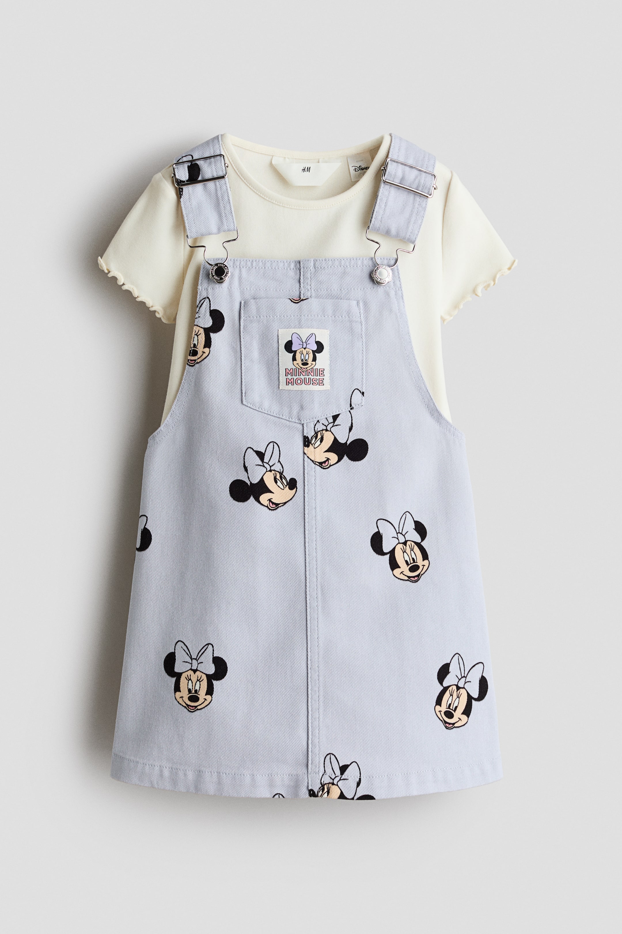 View larger image: A light blue overall dress, adorned with an all-over Minnie Mouse head print, is displayed from the front, layered over a cream short-sleeved t-shirt. The overall dress features adjustable bib straps with silver-toned buckles and buttons, and a chest pocket displaying a "Minnie Mouse" fabric label; the t-shirt showcases ruffled edges on its short sleeves.