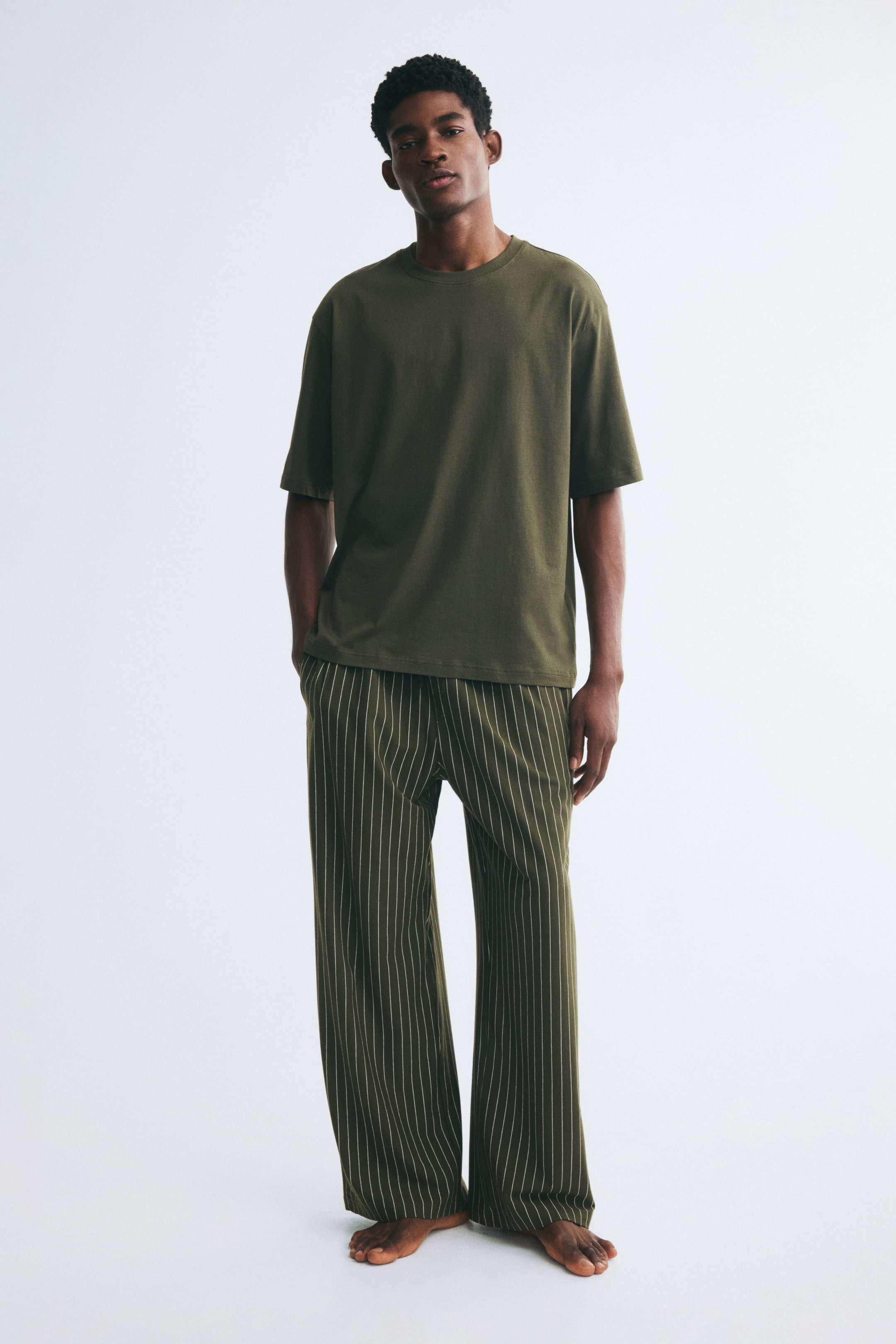 View larger image: A relaxed-fit pyjama set, featuring a solid olive-green T-shirt with a crew neck and matching pinstriped straight-leg pants, is being worn by the model. The model is standing with one hand resting in the side pocket of the pants.