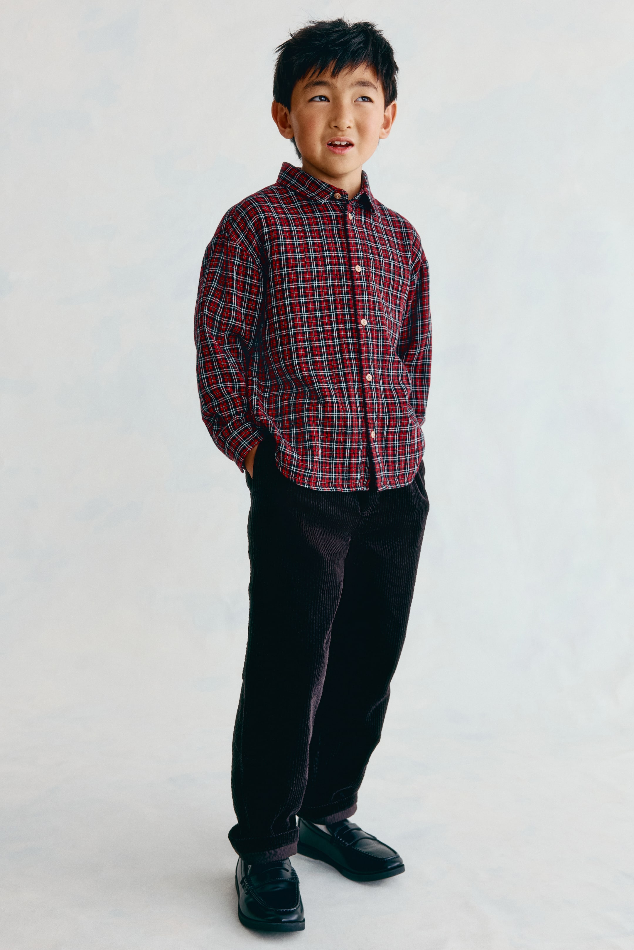 Voir une image plus grande: Ce pantalon en velours côtelé brun foncé H&M Adorables est présenté de face et se distingue par sa coupe droite et sa texture côtelée bien visible. Le pantalon est agencé avec une chemise boutonnée à carreaux rouges et des chaussures plates noires, tandis qu’une main repose dans une poche latérale.