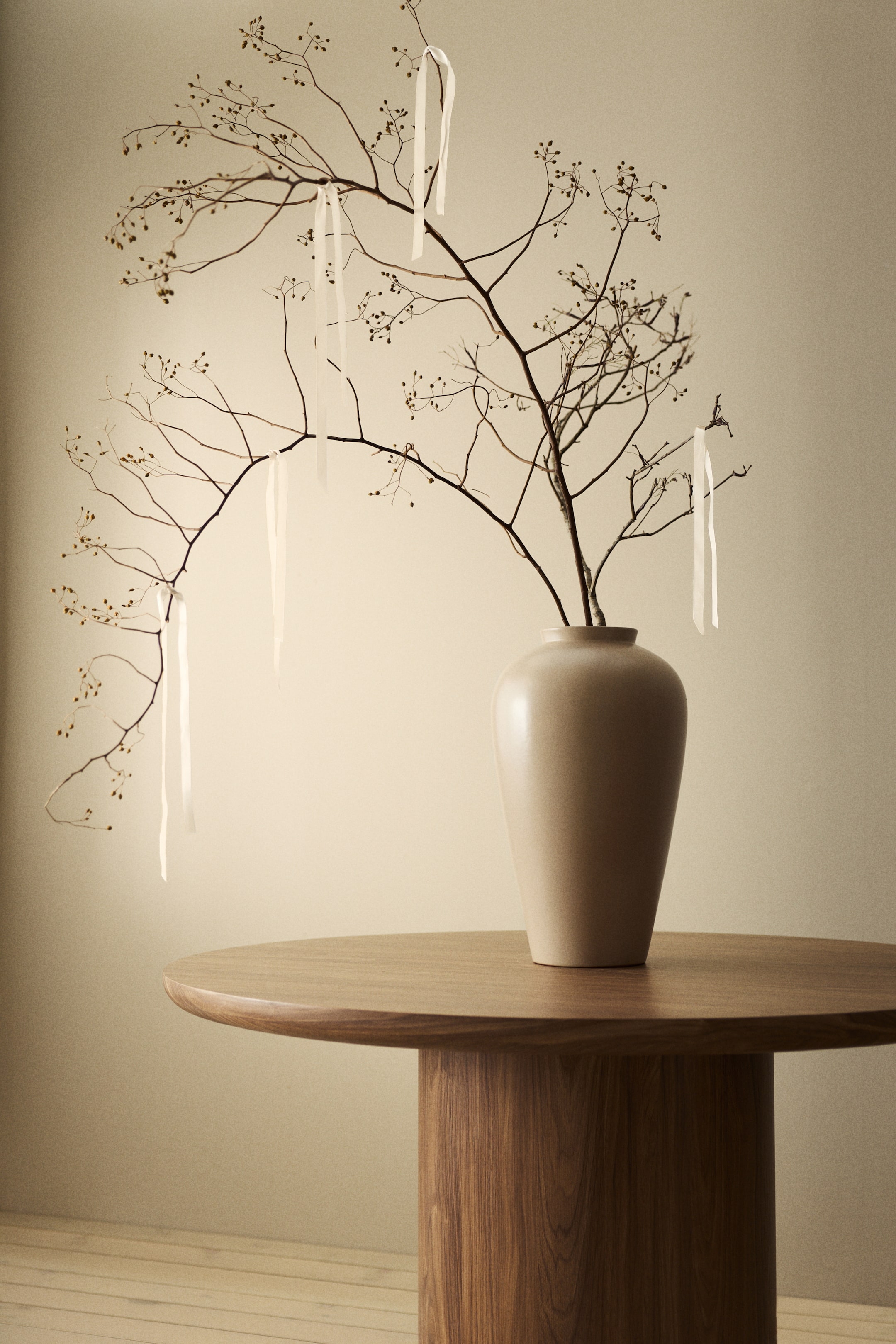 Agrandir l'image: Un vase en céramique beige clair est posé sur une table en bois, sa forme effilée visible sous un léger angle droit. Le vase présente une finition lisse et mate, s'élargissant de sa base jusqu'à un corps plein avant de se rétrécir au niveau du col, et il contient des branches séchées décorées de rubans blancs.