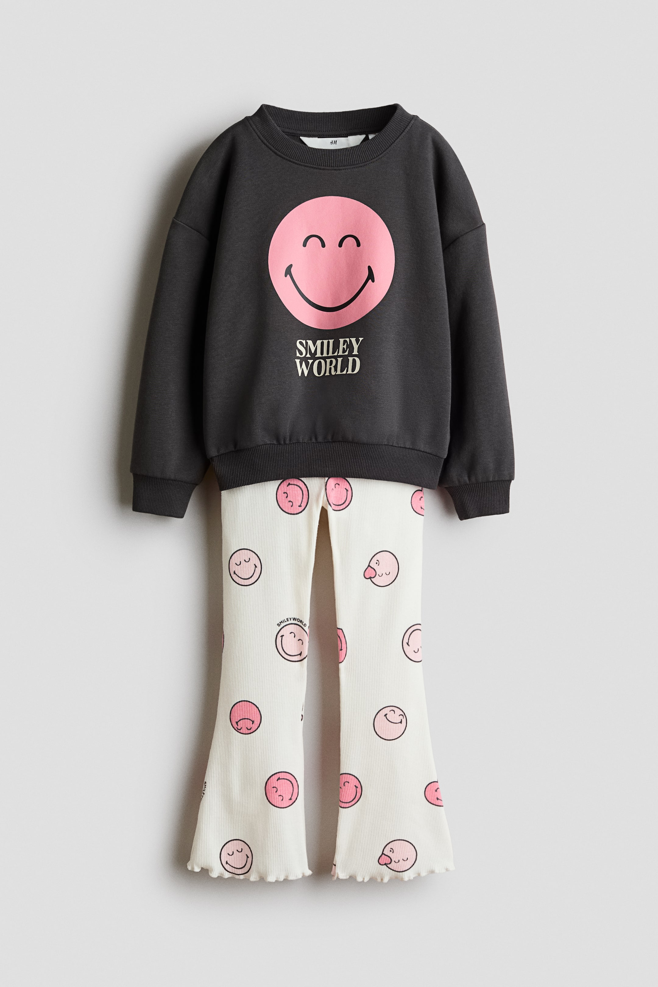 View larger image: The two-piece set showcases a dark gray crew-neck sweatshirt with ribbed cuffs and hem, adorned with a large pink smiley face graphic and "SMILEY WORLD" text on the front. Complementing off-white ribbed pants feature a flared leg design with ruffled hems and an all-over pattern of pink smiley faces.