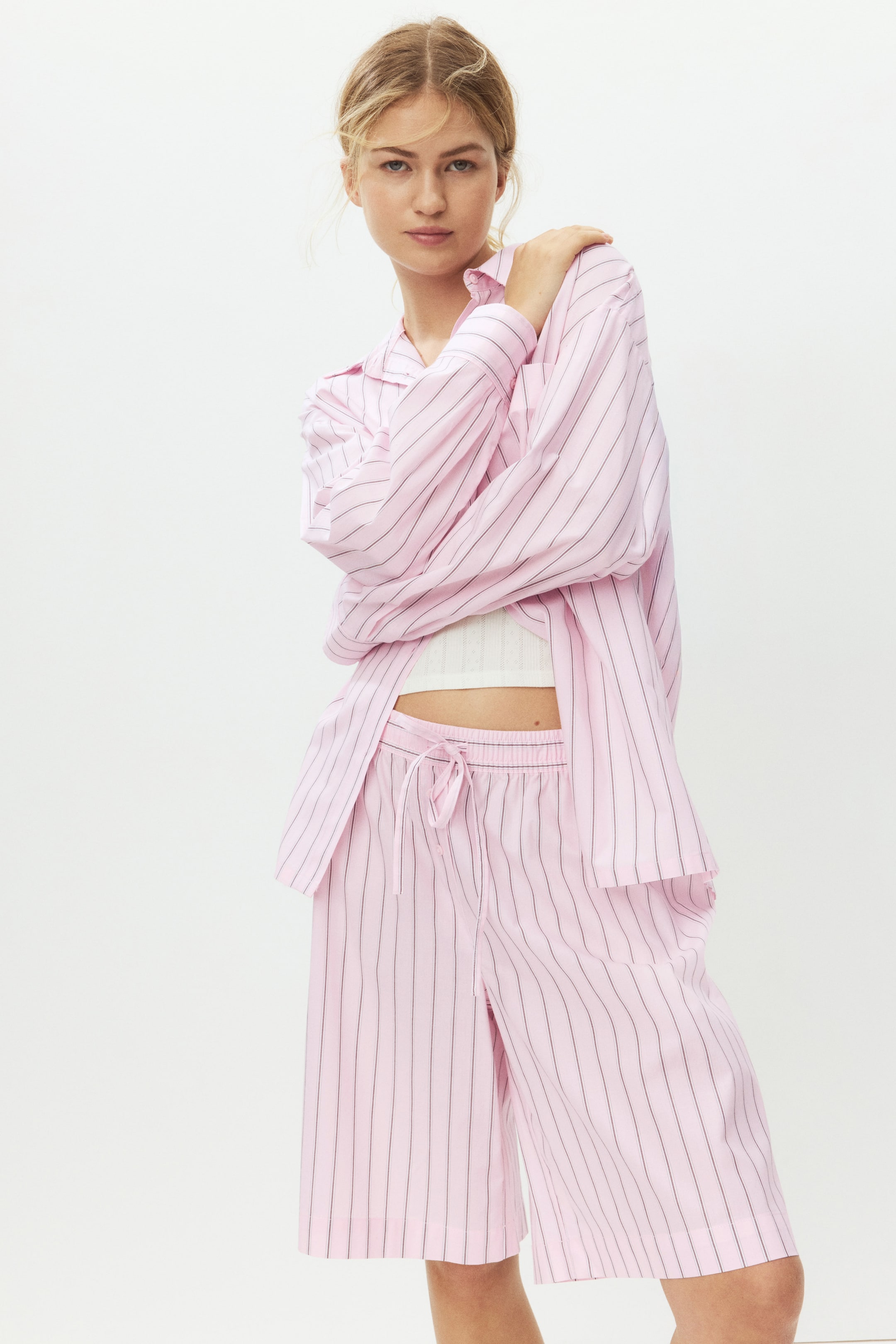 Agrandir l'image: Un ensemble de pyjama rose à rayures verticales noires est présenté, comprenant une chemise à col ouvert avec des manches longues superposée sur un haut côtelé blanc, et un short long jusqu'aux genoux avec une taille à cordon de serrage.