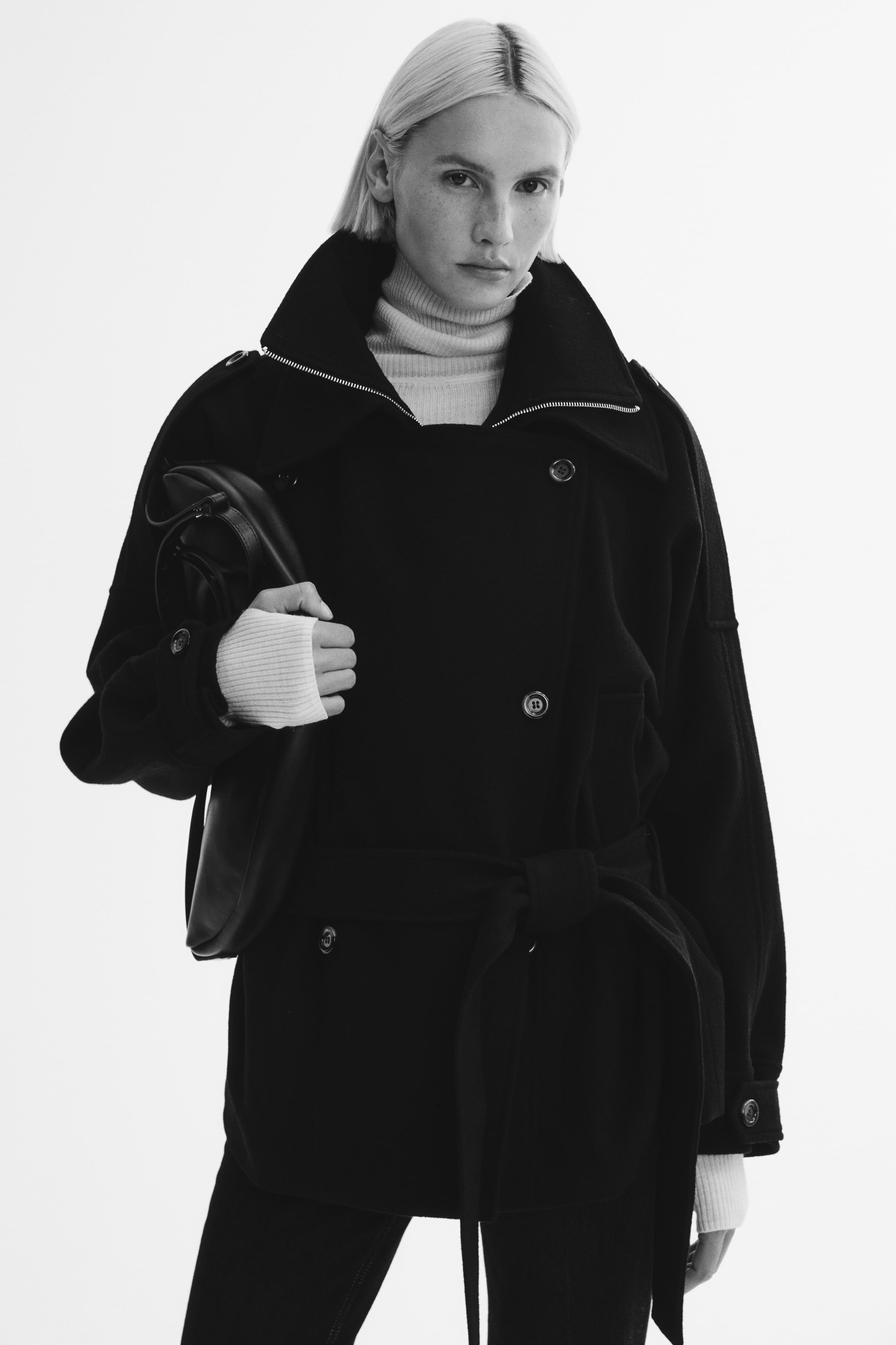 Ver imagen más grande: La modelo lleva un abrigo corto, negro y de doble botonadura, de corte oversize, sobre un jersey de cuello alto de canalé en color claro. El abrigo presenta un cuello doble con cuello cisterna interior con cremallera, un amplio cuello exterior, tiras abotonadas en los hombros y un cinturón a juego en la cintura. La modelo también lleva un bolso de piel negro bajo el brazo.