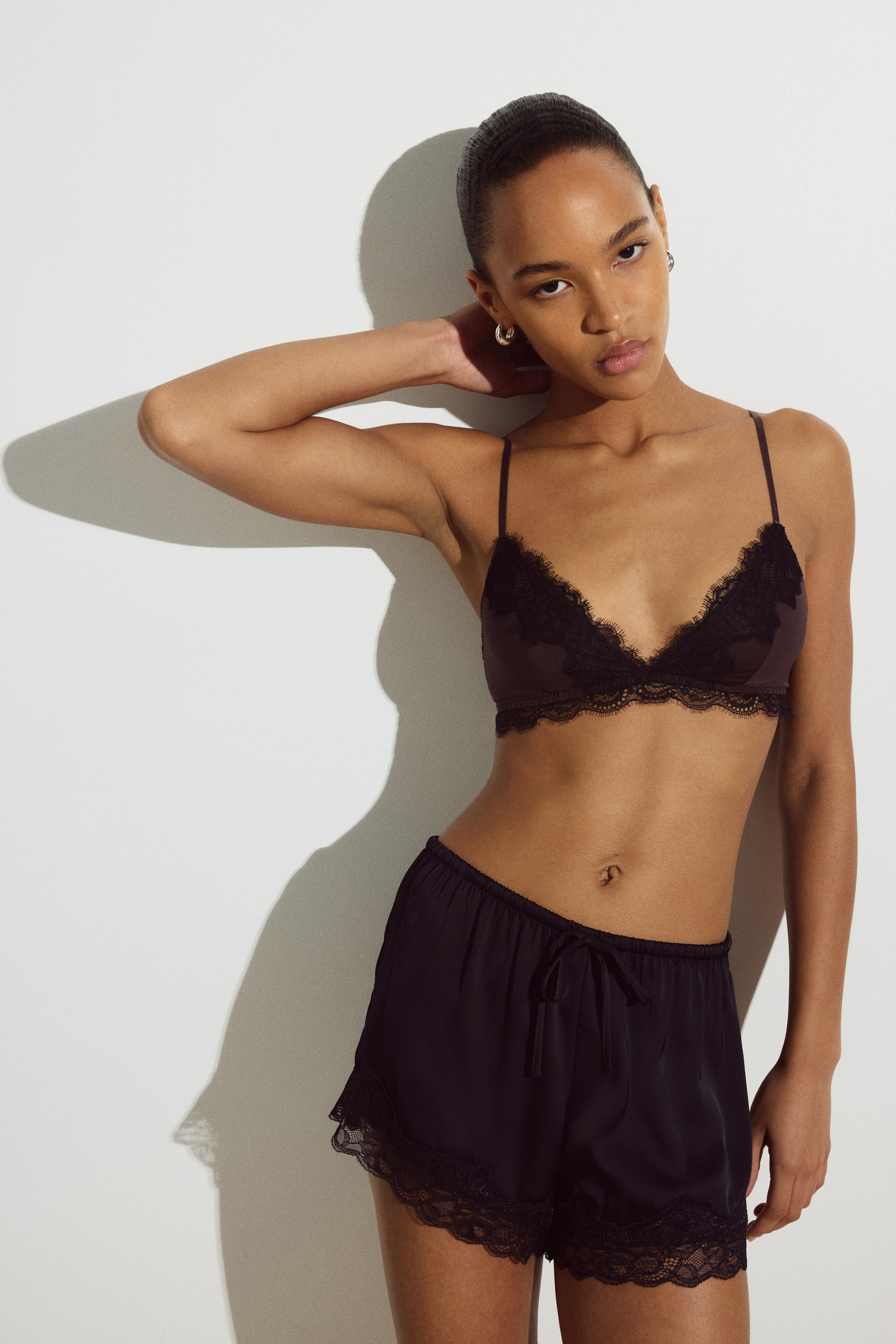 View larger image: The model is wearing a dark brown soft bra featuring microfiber cups with black lace trim along the top edges and band, along with thin, adjustable shoulder straps. The model is showcasing the bra while wearing matching dark shorts with black lace trim.