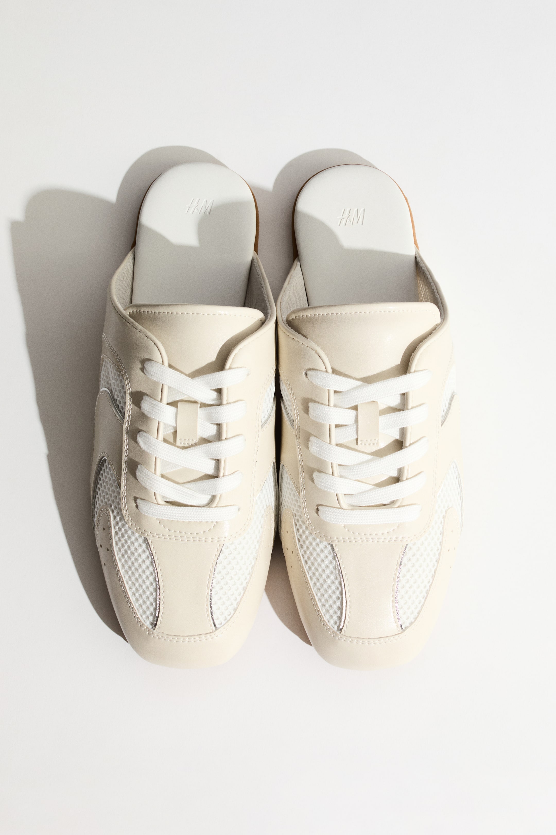 Ingrandisci l'immagine: Mules stile sneaker color avorio con lacci bianchi, inserti in mesh sui lati e sulla punta, e sezioni lisce color crema che formano la struttura principale. Sono posizionate una accanto all'altra, rivolte in avanti, con il logo H&M impresso su ogni soletta bianca.