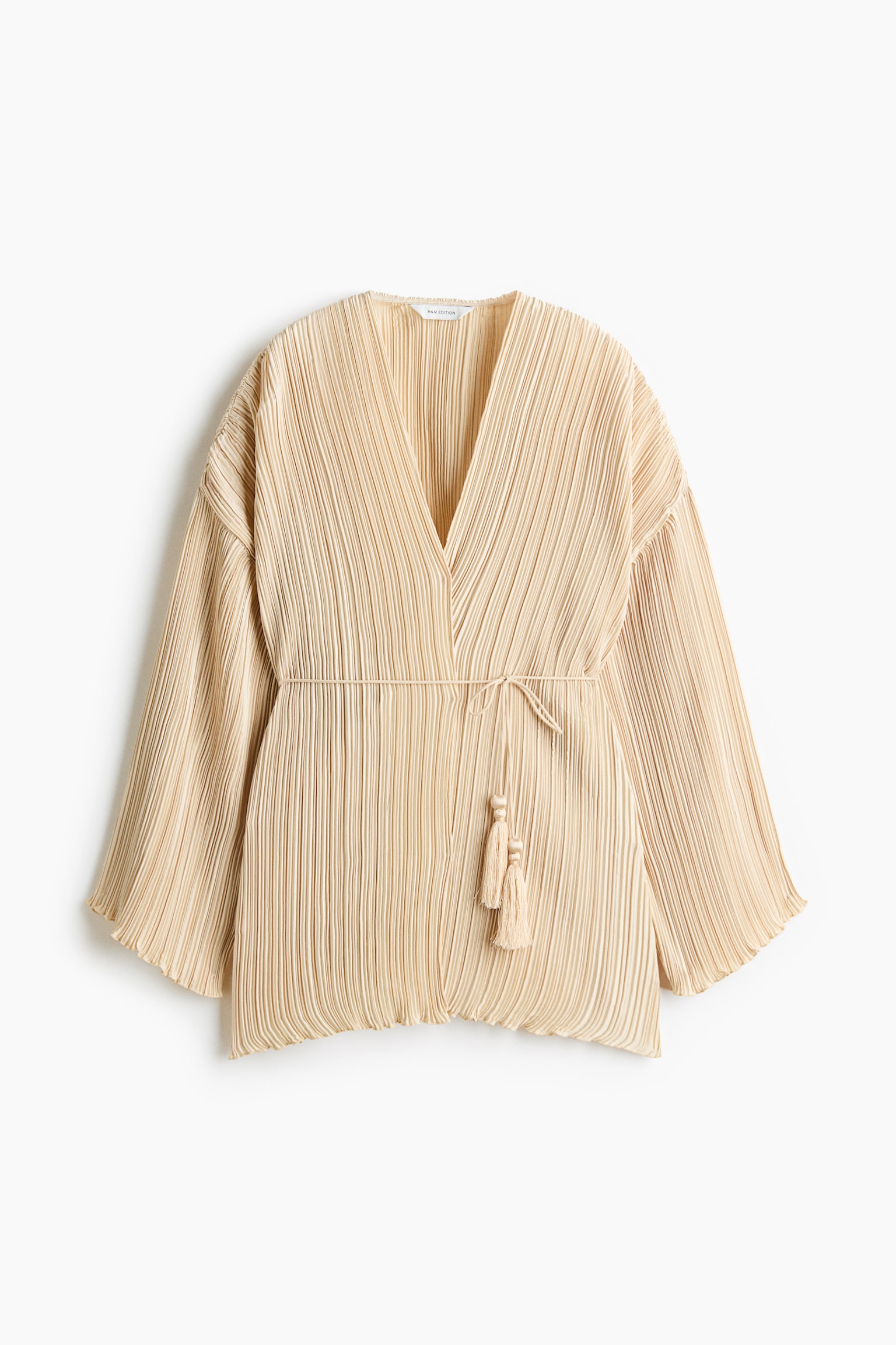 A beige plissÃ© wrap top is shown front-on, featuring a deep V-neckline, long wide sleeves, and fine vertical pleating throughout. A thin matching tie belt cinches in the waist, with two decorative beaded tassels hanging down.