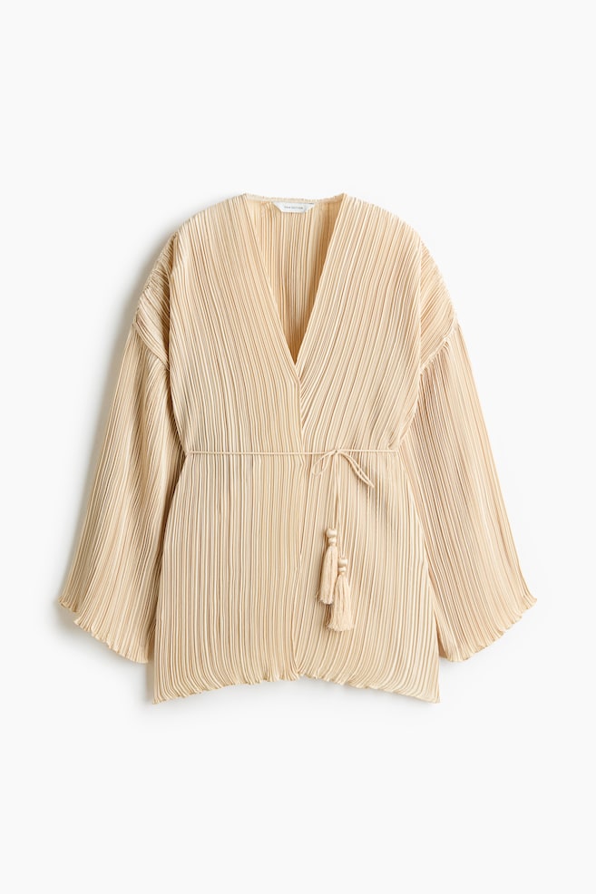 Un haut portefeuille plissÃ© beige est prÃ©sentÃ© de face, avec un profond col en V, de longues manches larges et de fines plissures verticales sur lâensemble du modÃ¨le. Une fine ceinture assortie Ã  nouer marque la taille, avec deux pompons dÃ©coratifs perlÃ©s qui pendent. - 1