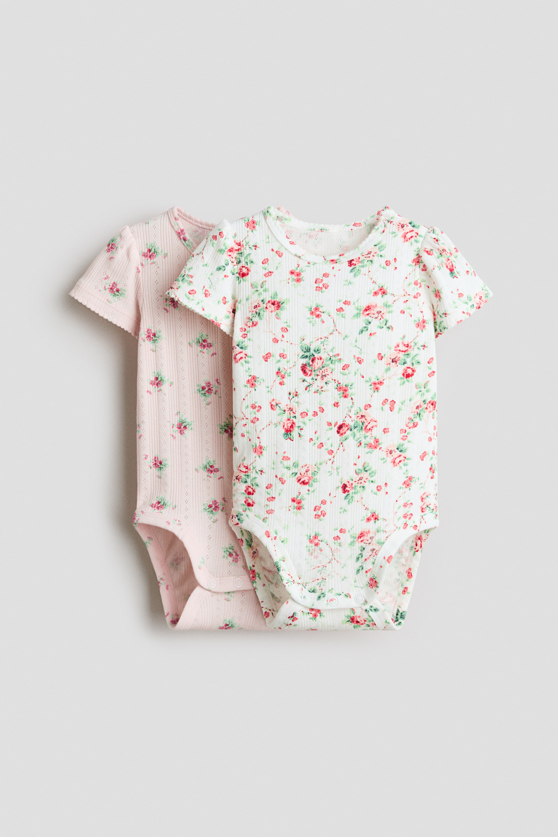Größeres Bild anzeigen: Zwei kurzärmelige Baby-Bodys präsentieren eine gerippte Strickstruktur und Druckknopfverschlüsse im Schritt, in einer Frontalansicht dargestellt. Der hellrosa Body zeigt ein dezentes florales Pointelle-Muster und gesäumte Kanten an den kurzen Ärmeln. Der cremeweiße Body weist einen auffälligen Allover-Blumendruck mit roten Rosen und grünen Blättern auf.