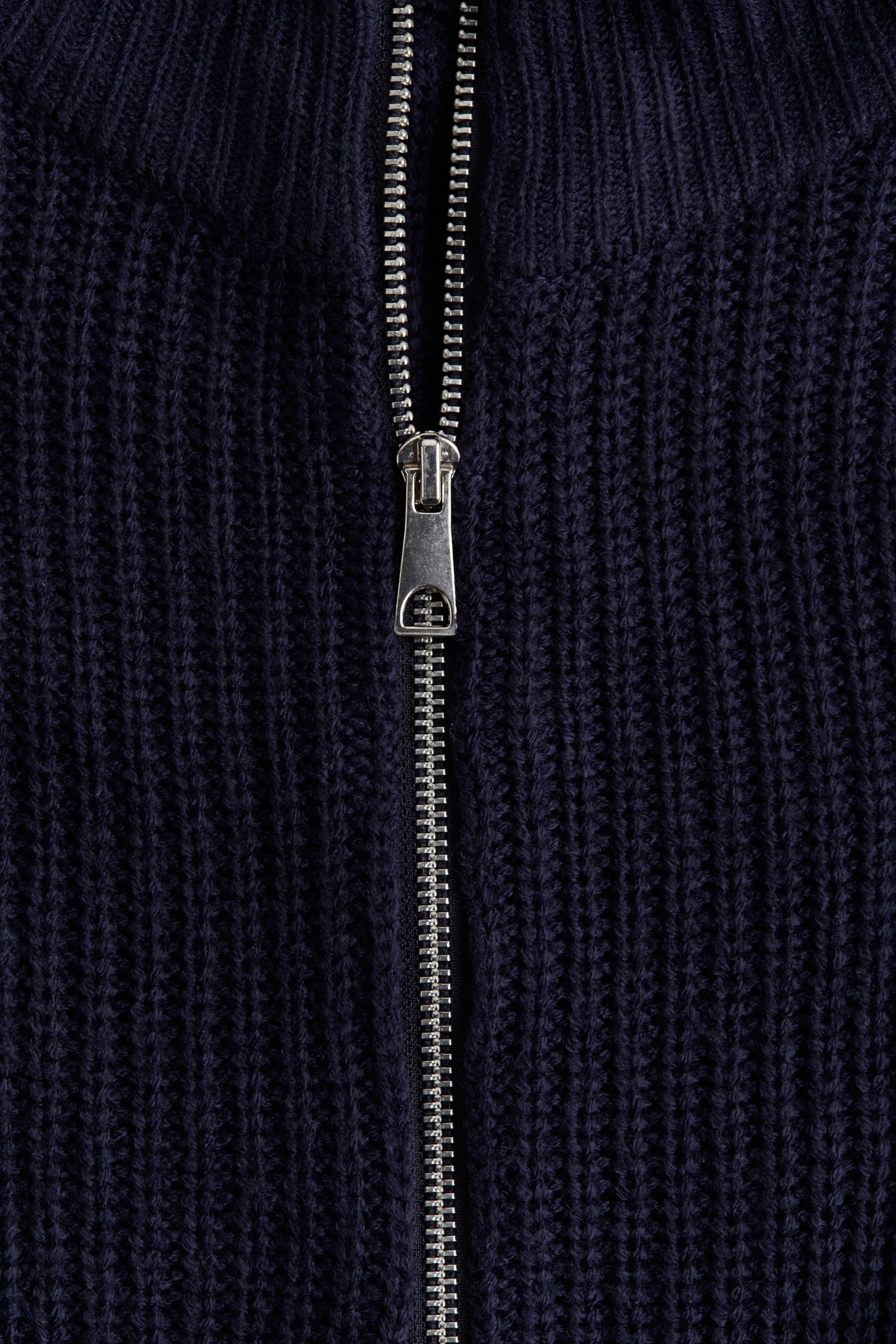 View larger image: A dark blue chunky knit jumper is displaying a silver-toned metal zipper running down its center front, with the zipper pull visible.