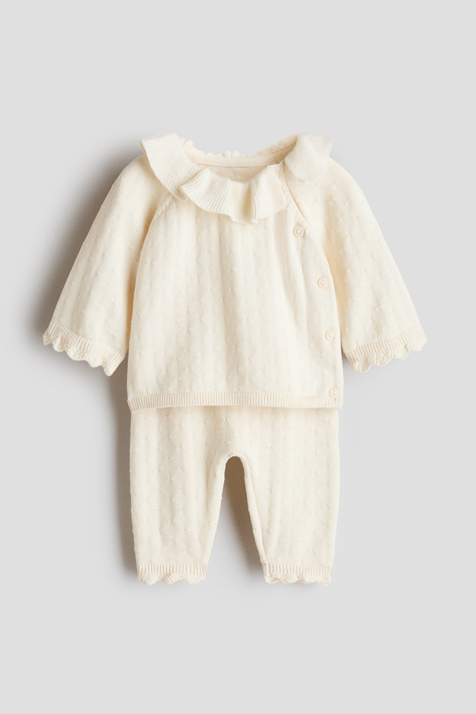 This cream-coloured baby set in soft, pointelle-knit cotton is shown from the front, featuring a long-sleeve cardigan with a ribbed, flounced collar, an asymmetrical side-button closure, and scalloped cuffs. The matching trousers are displayed below, showing a tapered fit with rib-trimmed, scalloped ankle hems.