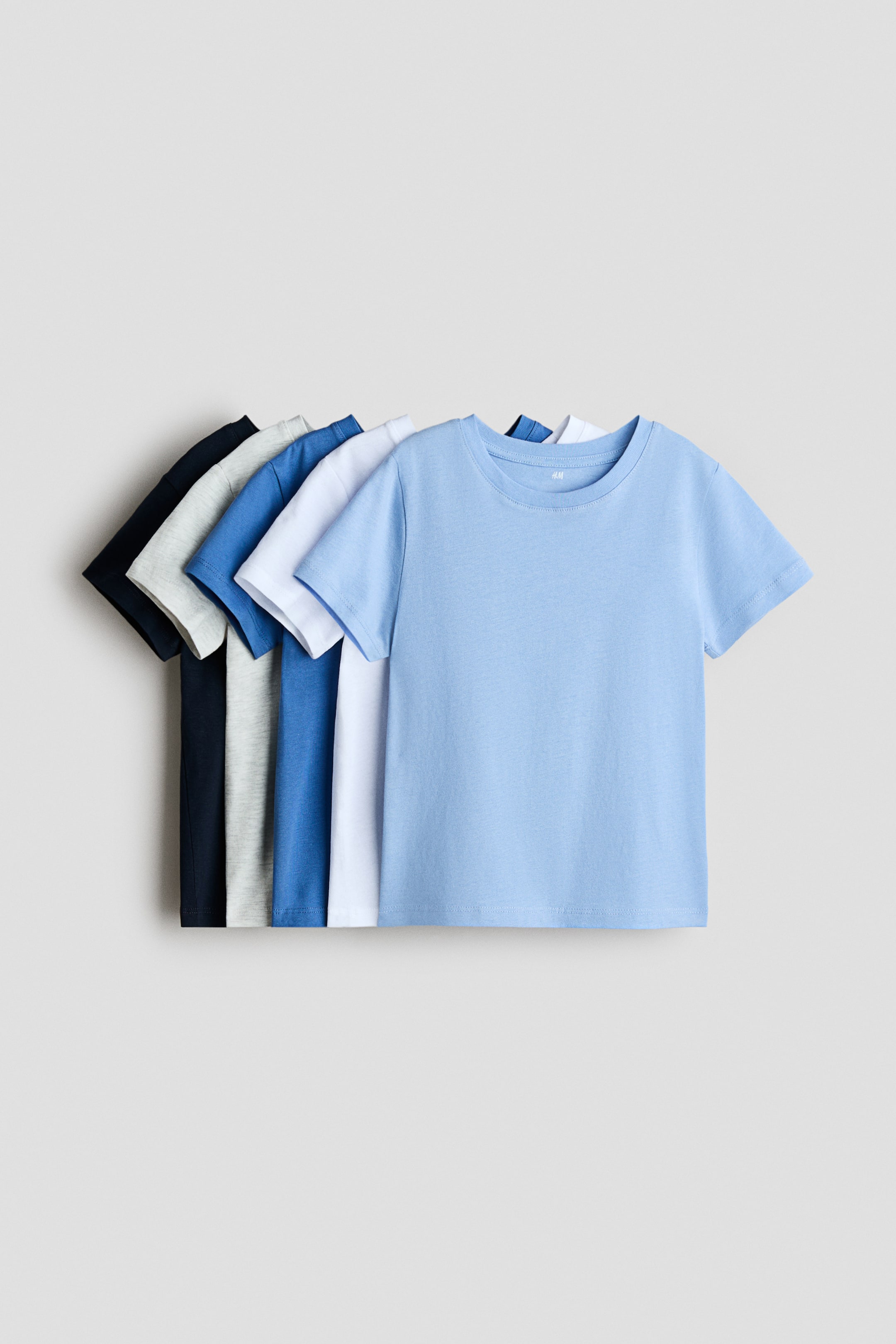 View larger image: Five soft cotton jersey t-shirts are arranged with their fronts visible, spread out horizontally. The leading t-shirt is in a light blue shade, showcasing a round neckline, short sleeves, and a straight-cut hem. Behind it, other t-shirts can be seen in white, medium blue, light grey, and dark blue.