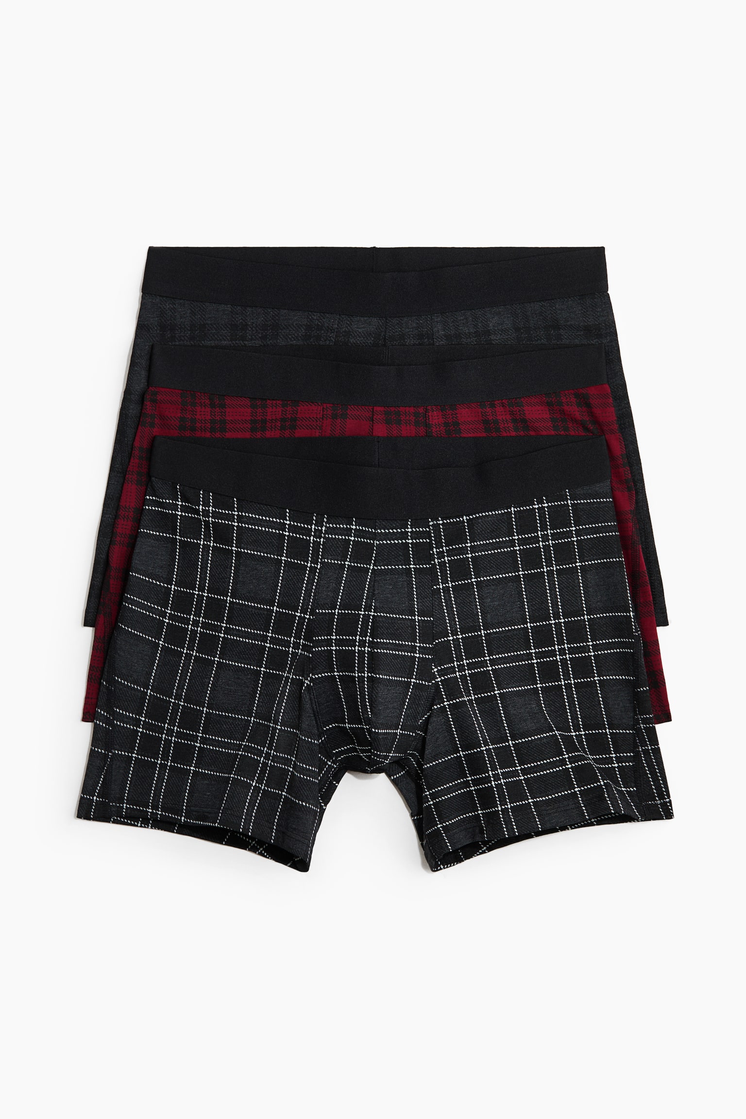 A three-pack of men's boxer briefs is laid flat and facing forward in a layered stack, with each pair featuring a solid black elastic waistband. The front pair displays a dark grey and white windowpane plaid pattern, the middle pair shows a red and black plaid pattern, and the back pair shows a dark grey plaid pattern.