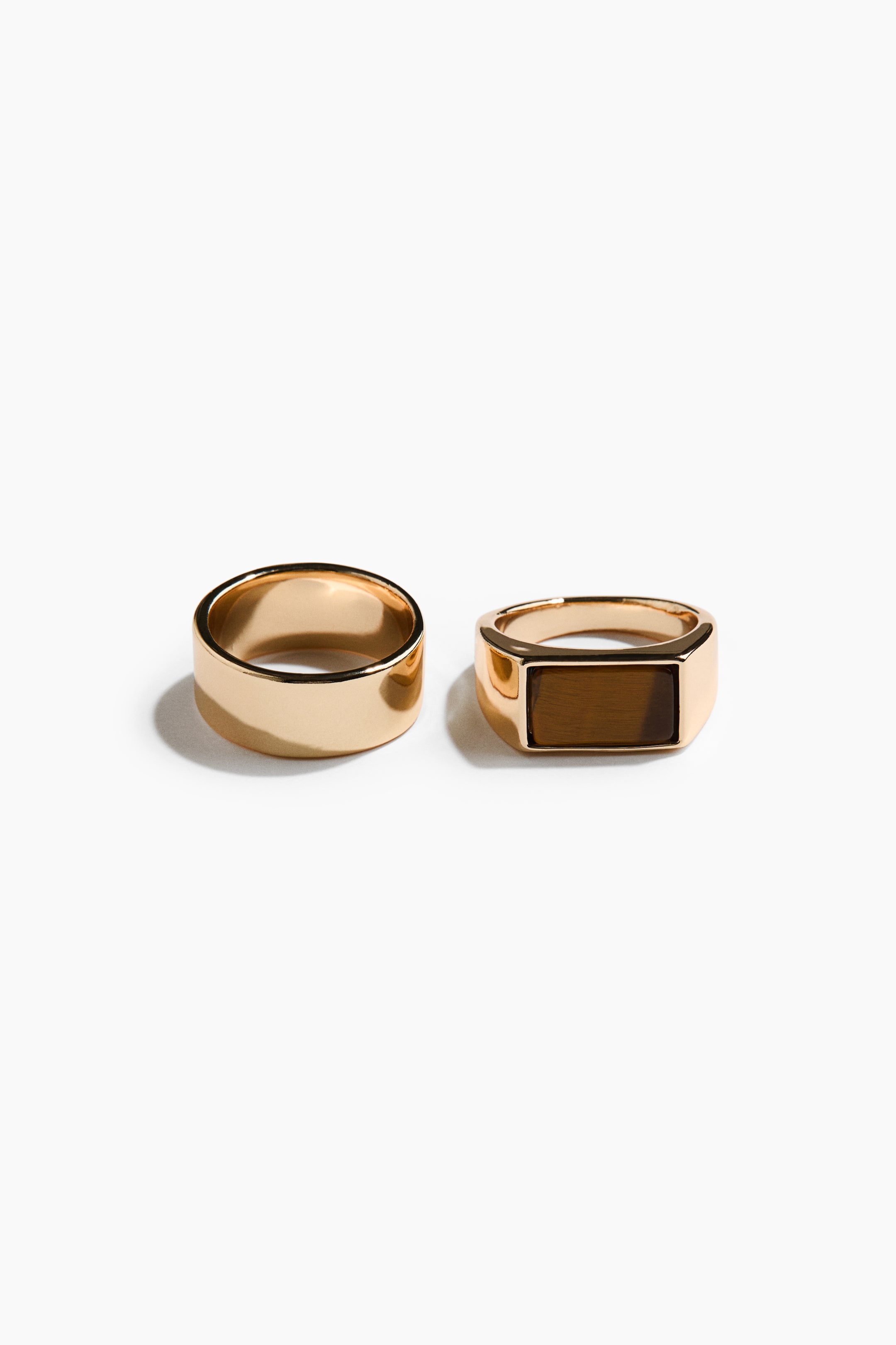 View larger image: Two rings are on display: a wide, gold-toned band with a smooth, polished finish, and a gold-toned signet ring featuring a rectangular face with a brown, striated inlay.