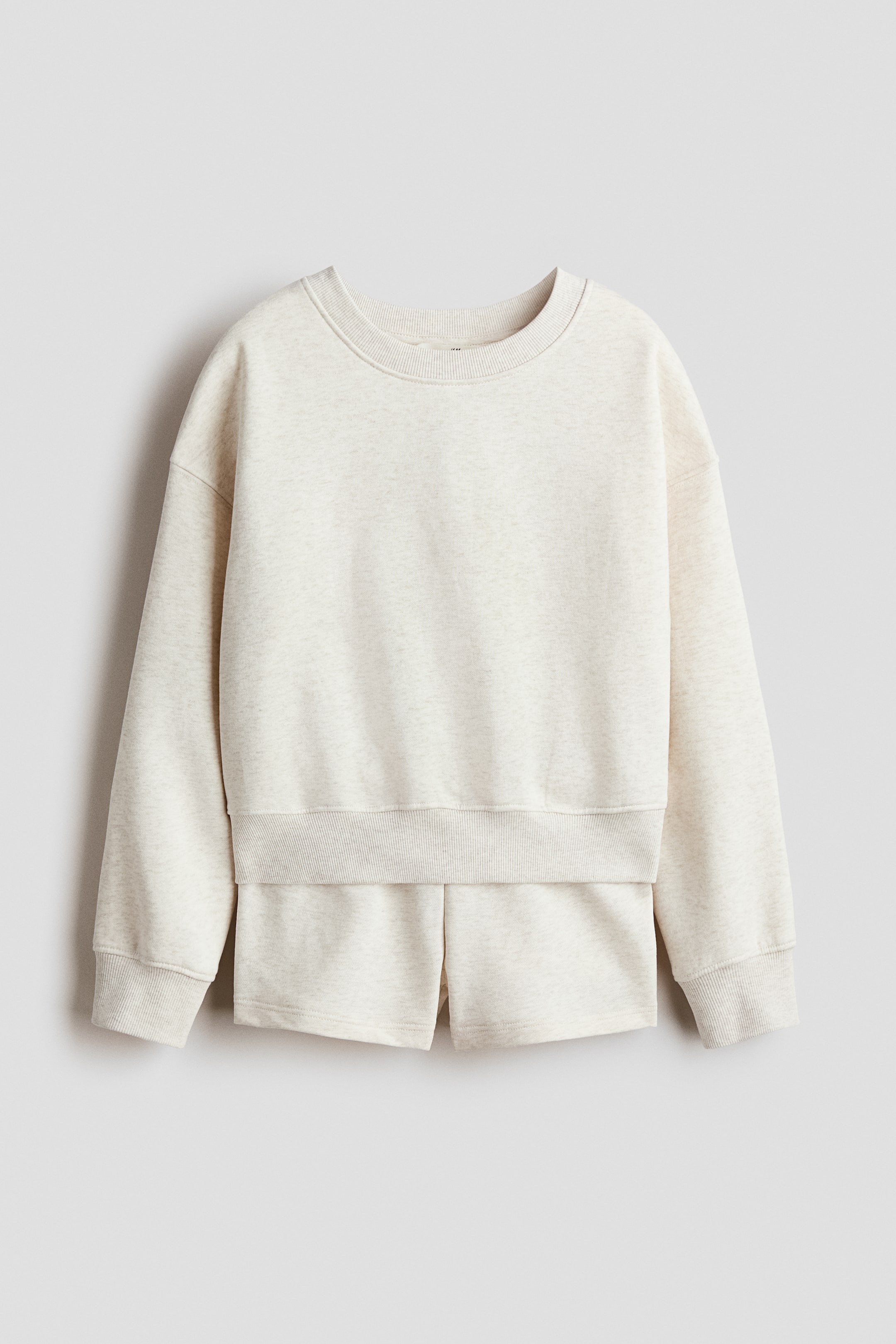 Größeres Bild anzeigen: Ein hellbeiges Sweatshirt-Set wird von vorne präsentiert, bestehend aus einem langärmeligen Oberteil mit geripptem Rundhalsausschnitt, überschnittenen Schultern, gerippten Bündchen und einem gerippten Saum, zusammen mit passenden Shorts, die unter dem Oberteil sichtbar sind und Schlitze an den Säumen zeigen.