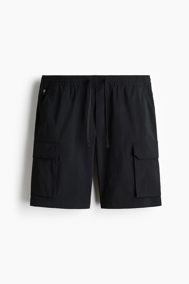 Black nylon cargo shorts are showcased from the front, featuring an elasticated, drawstring waist and a faux fly. Diagonal side pockets are visible, with an internal zipped pocket appearing on the left side, while flap leg pockets with concealed press-studs extend down each leg, contributing to a regular fit. - 1