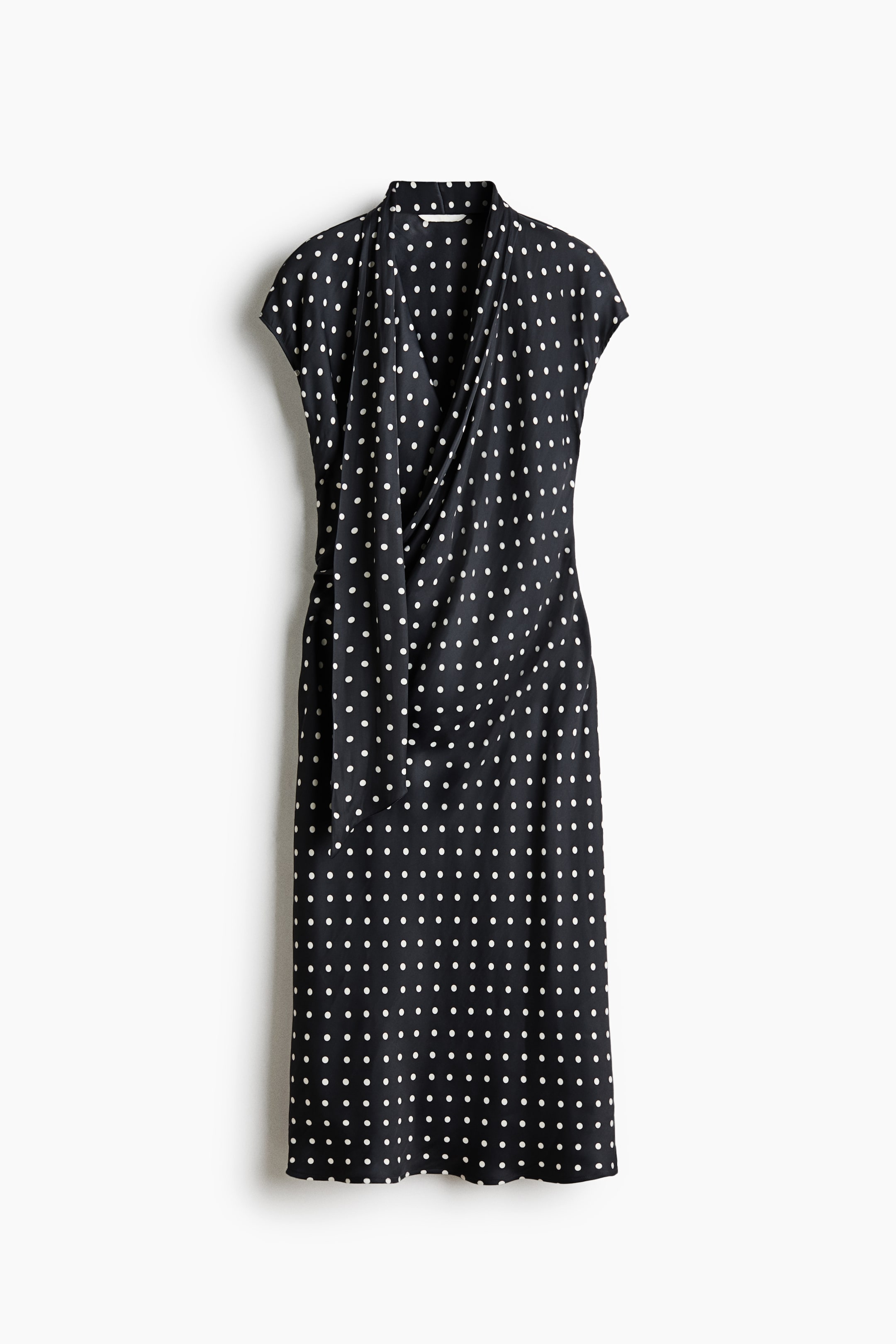 View larger image: A black midi dress with white polka dots features a draped cowl neckline and short cap sleeves. The wrap-front design creates a draped effect across the bodice, and a self-tie detail is visible on the left side of the waist.