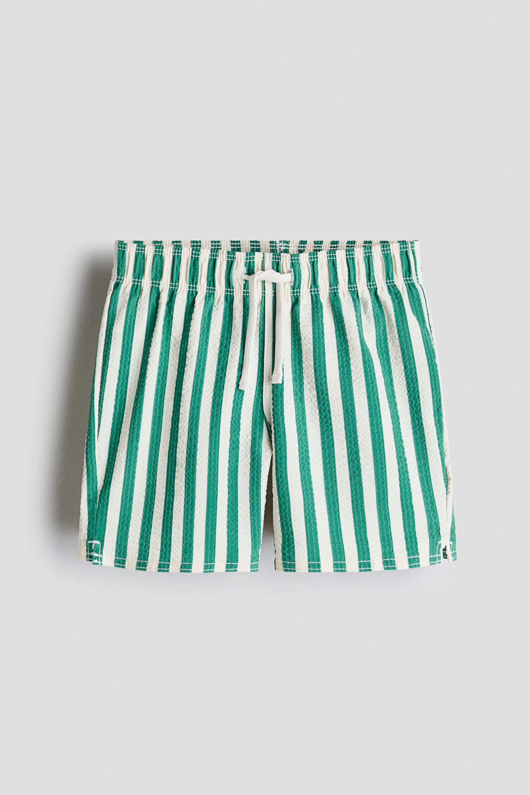 View larger image: H&M Adorables swim shorts showcase their front, featuring a textured fabric with alternating green and off-white vertical stripes. They are designed with an elastic waistband, a white drawstring, and display small slits on both side hems.