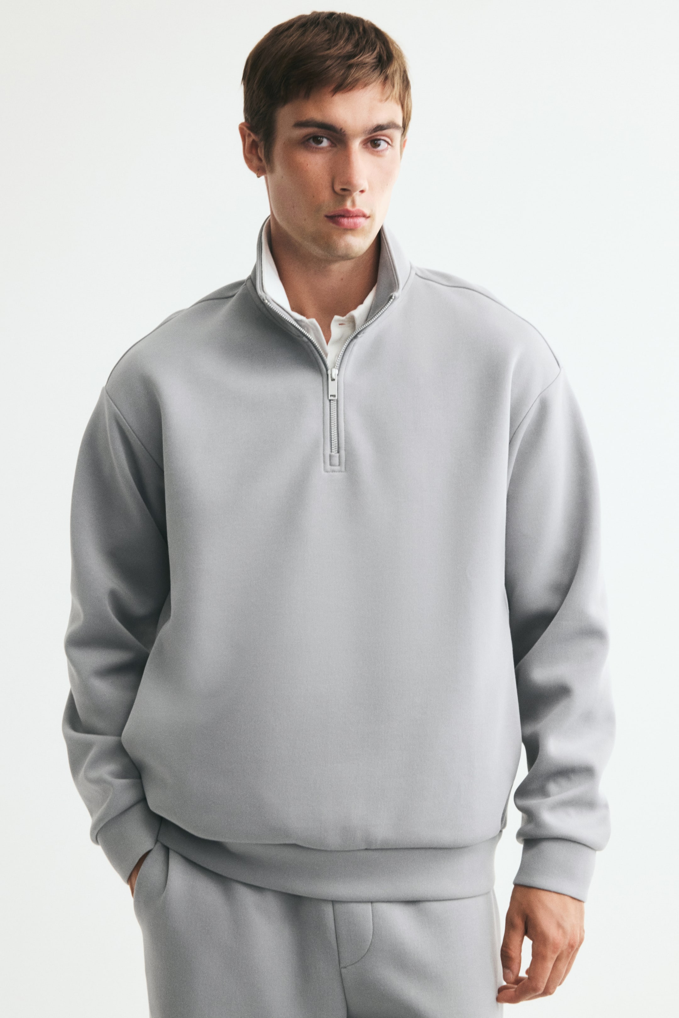 Voir une image plus grande: Un chandail gris pâle à demi-fermeture éclair, confectionné dans un tissu lisse de poids moyen, est porté par le mannequin par-dessus une chemise blanche à col. Le chandail à coupe décontractée présente un col montant, des épaules tombantes et des bordures côtelées, et il est agencé avec un pantalon de jogging gris assorti.