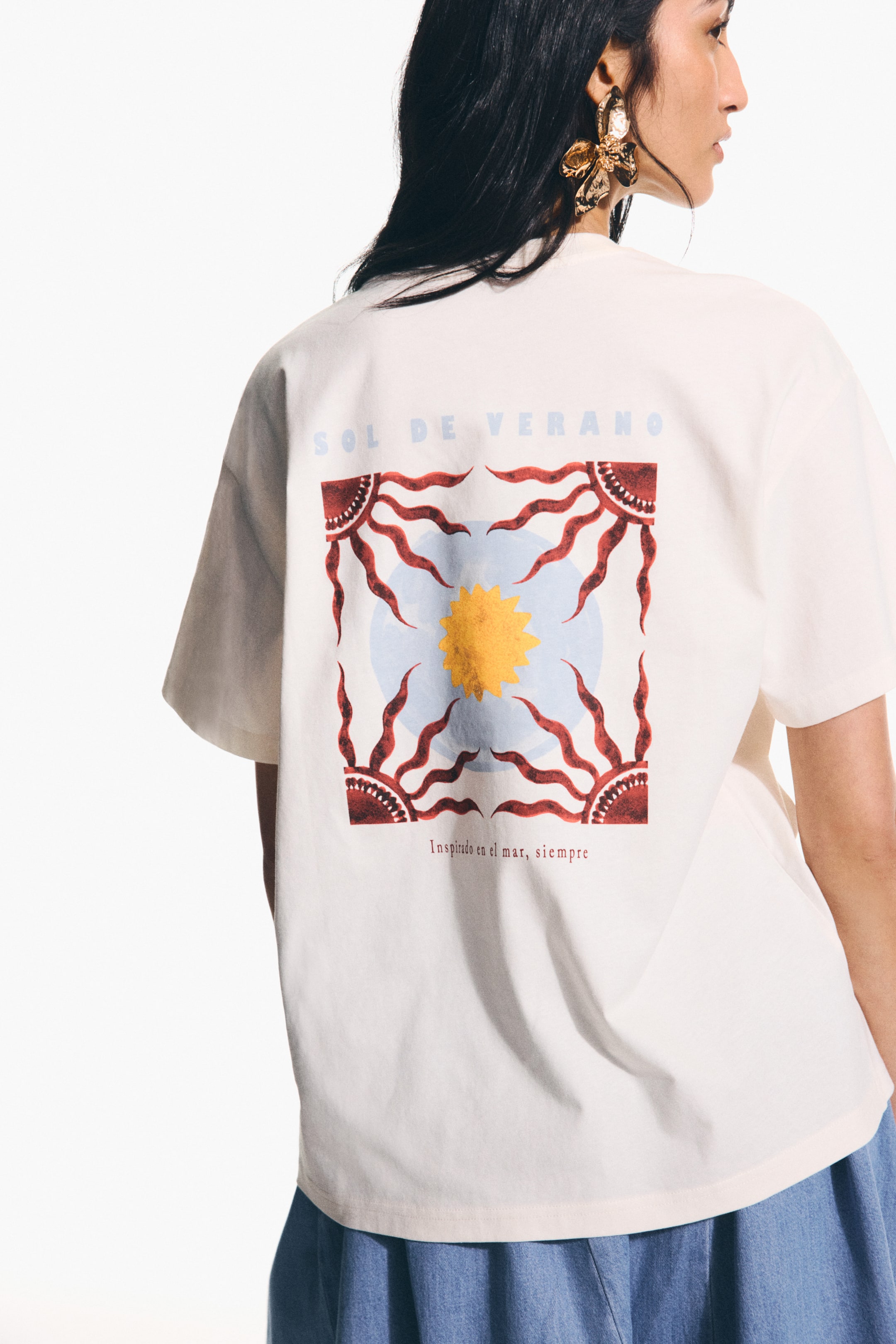 View larger image: An off-white t-shirt is being presented from the back, showcasing a large square graphic that displays 'SOL DE VERANO' at the top, a central yellow sun with red rays on a light blue background, and a border of four sun-like corners connected by wavy red lines. The text 'Inspirado en el mar, siempre' also appears at the bottom of this graphic.