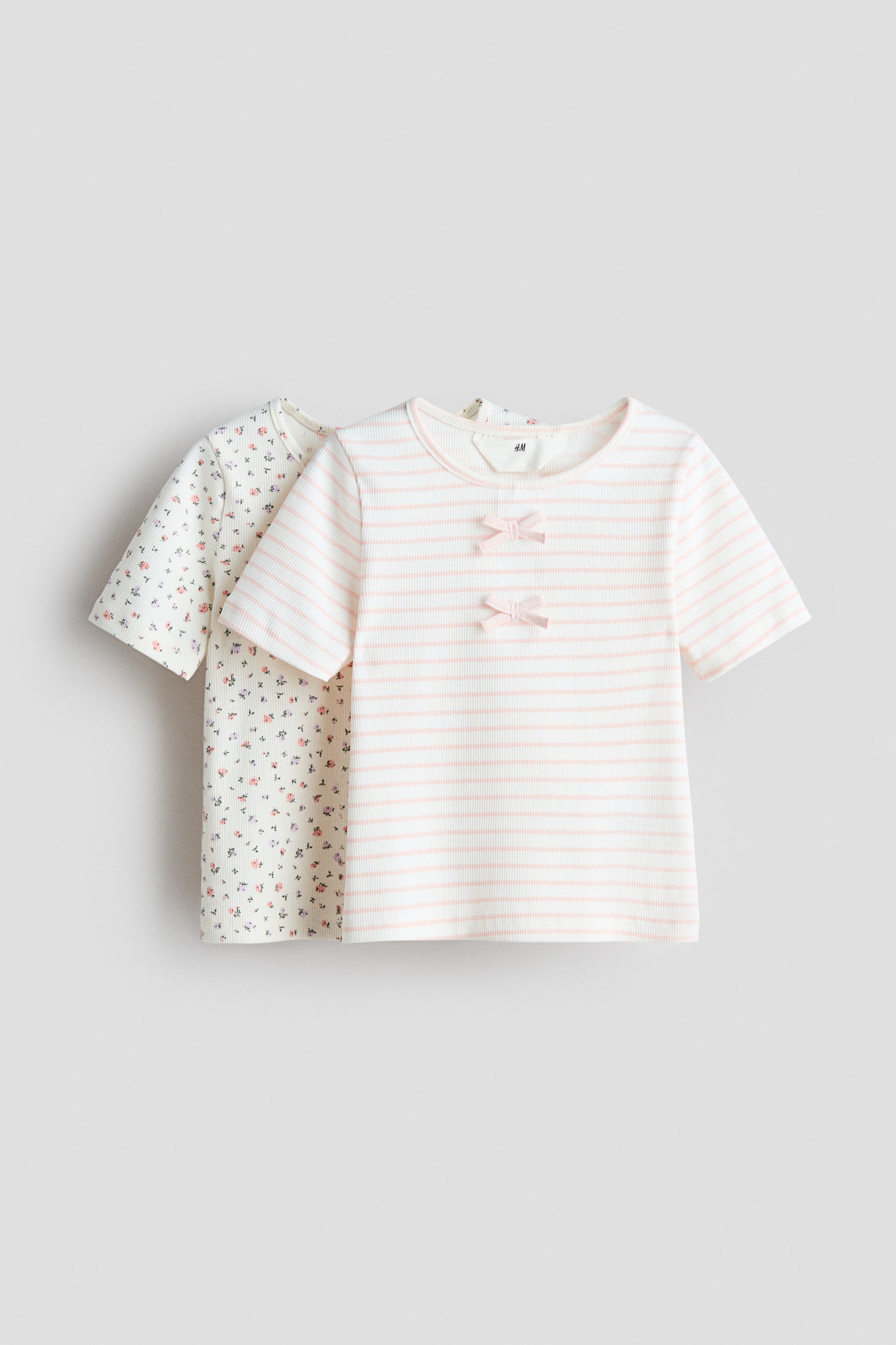 View larger image: Two short-sleeved tops are showcased at the FRONT. The front top boasts an off-white ribbed knit with horizontal light pink stripes, accentuated by two small pink bows on the bodice. The layered top behind features an off-white ribbed knit adorned with a delicate floral pattern.
