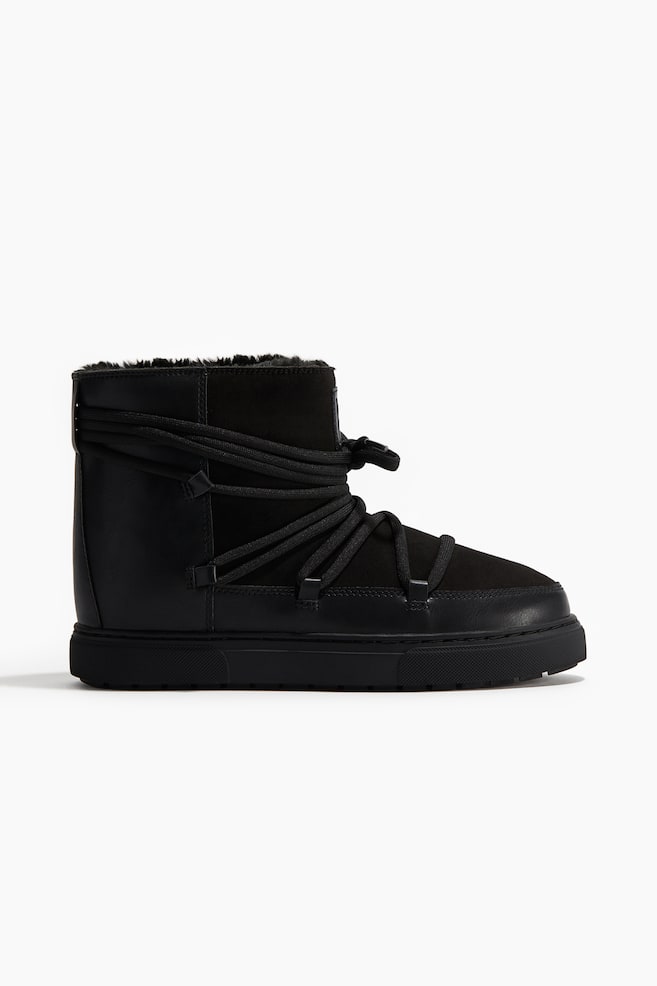 Une botte à la cheville noire et rembourrée présente une doublure duveteuse visible autour de son bord supérieur. Des lacets noirs épais s'enroulent horizontalement autour de la cheville et verticalement sur le devant du pied, fixés par de petites boucles sur les côtés, le tout reposant sur une semelle noire épaisse de 3 cm de haut avec un dessous cannelé. - 1