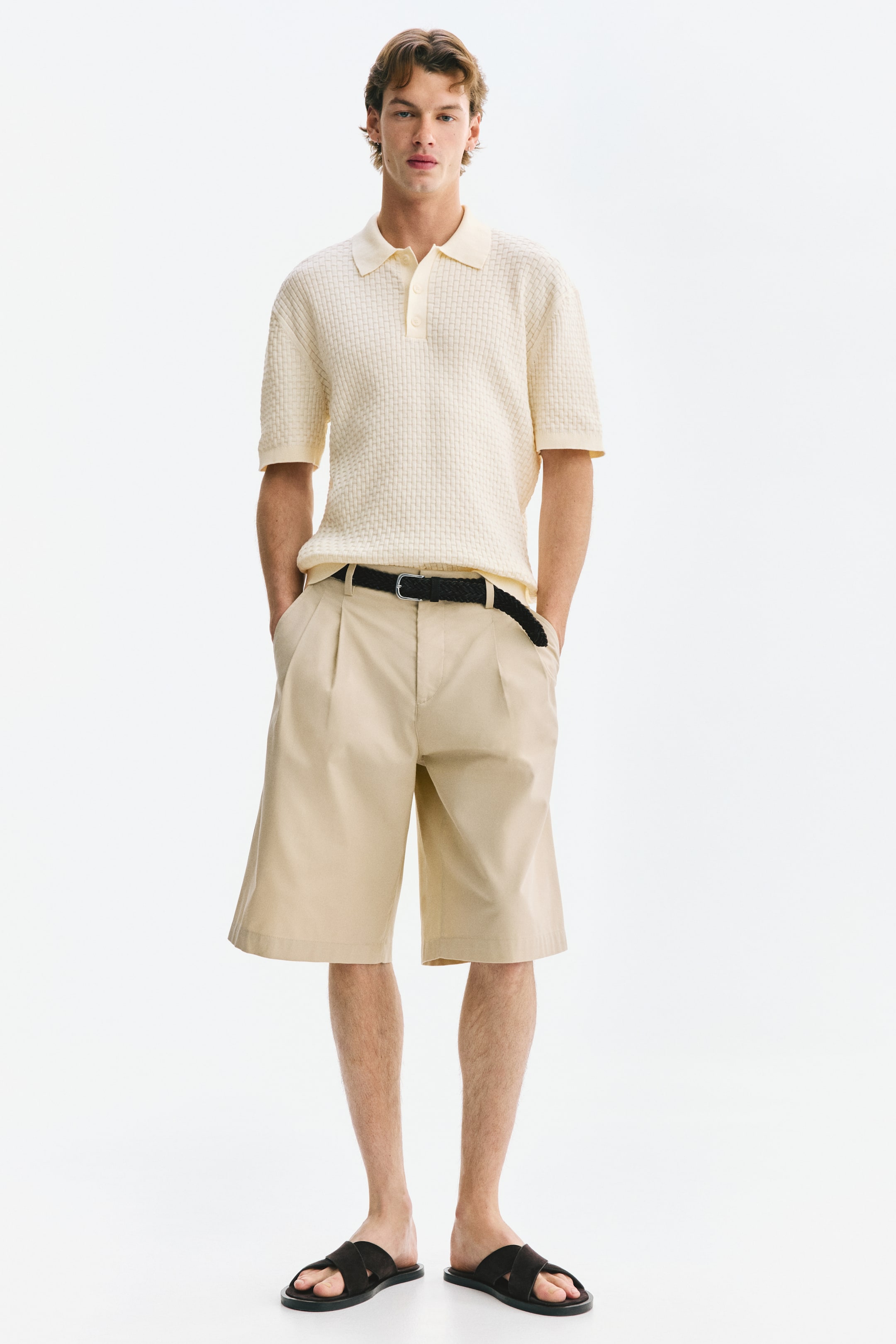 Ver imagen más grande: Una camisa polo de punto en color crema claro, que presenta un cuello, una tapeta de dos botones, mangas cortas con puños acanalados y un dobladillo acanalado, está siendo llevada por el modelo. El modelo la lleva metida dentro de unos pantalones cortos beige con pliegues, asegurados con un cinturón negro.