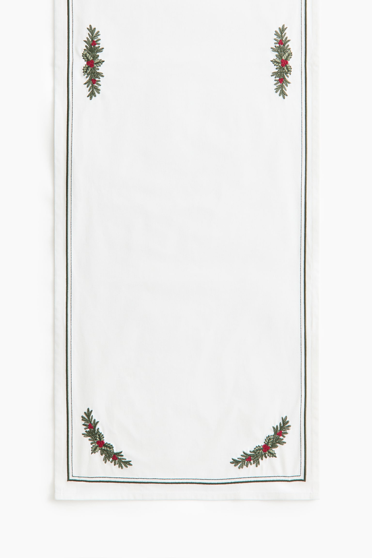 Un camino de mesa rectangular blanco cuelga en vertical y muestra la parte frontal con motivos bordados de ramas de pino verdes y bayas rojas en cada esquina. Un fino borde cosido en verde oscuro discurre paralelo a los bordes exteriores de la tela.