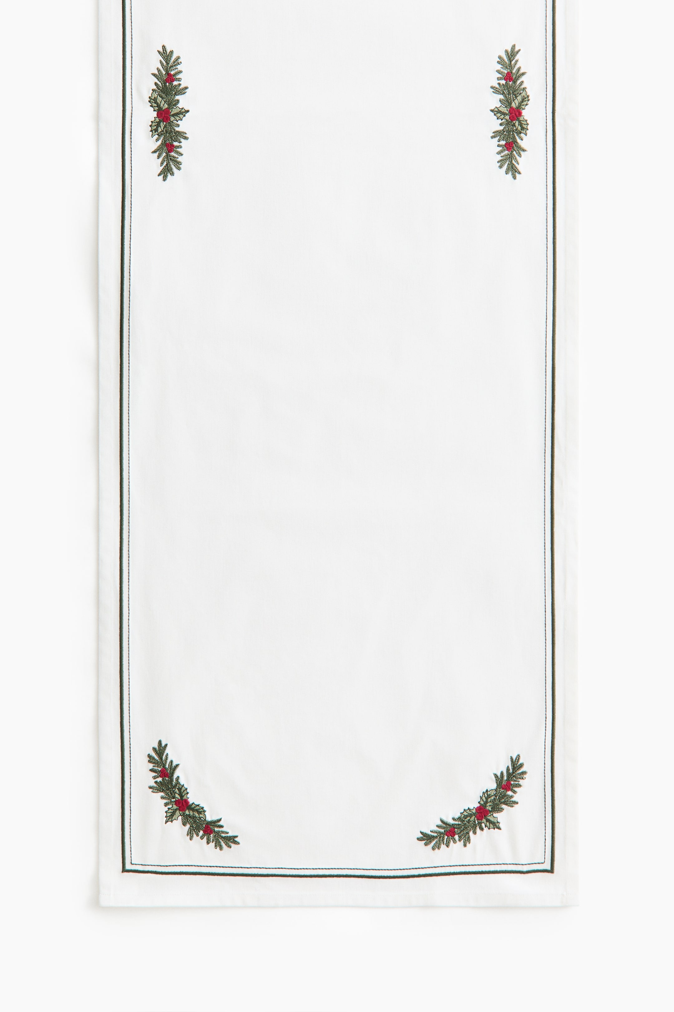 View larger image: A white rectangular table runner is hanging vertically, showing the front side with embroidered green pine branch and red berry motifs in each corner. A thin dark green stitched border runs parallel to the outer edges of the fabric.