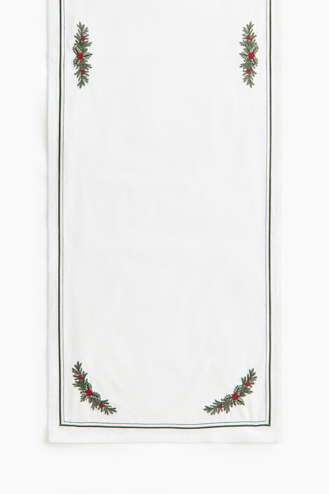 A white rectangular table runner hangs vertically, showing the front with embroidered green pine branch and red berry motifs in each corner. A thin, dark green stitched border runs parallel to the outer edges of the fabric. - 1