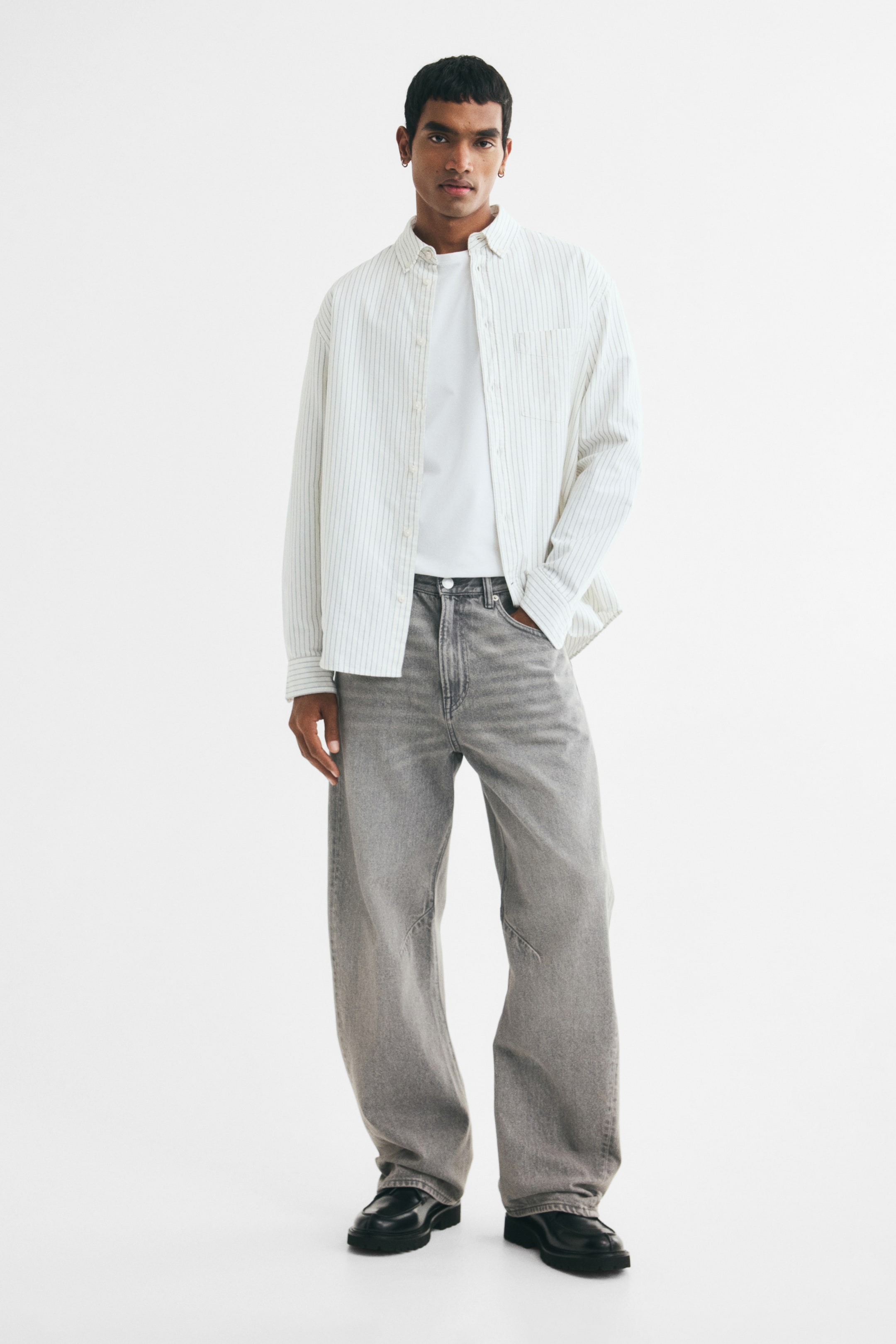 View larger image: Gray loose-fit jeans with a washed finish and wide legs are worn by the model. The model is placing one hand into a front pocket of the jeans, which are styled with a white T-shirt and an open striped shirt.