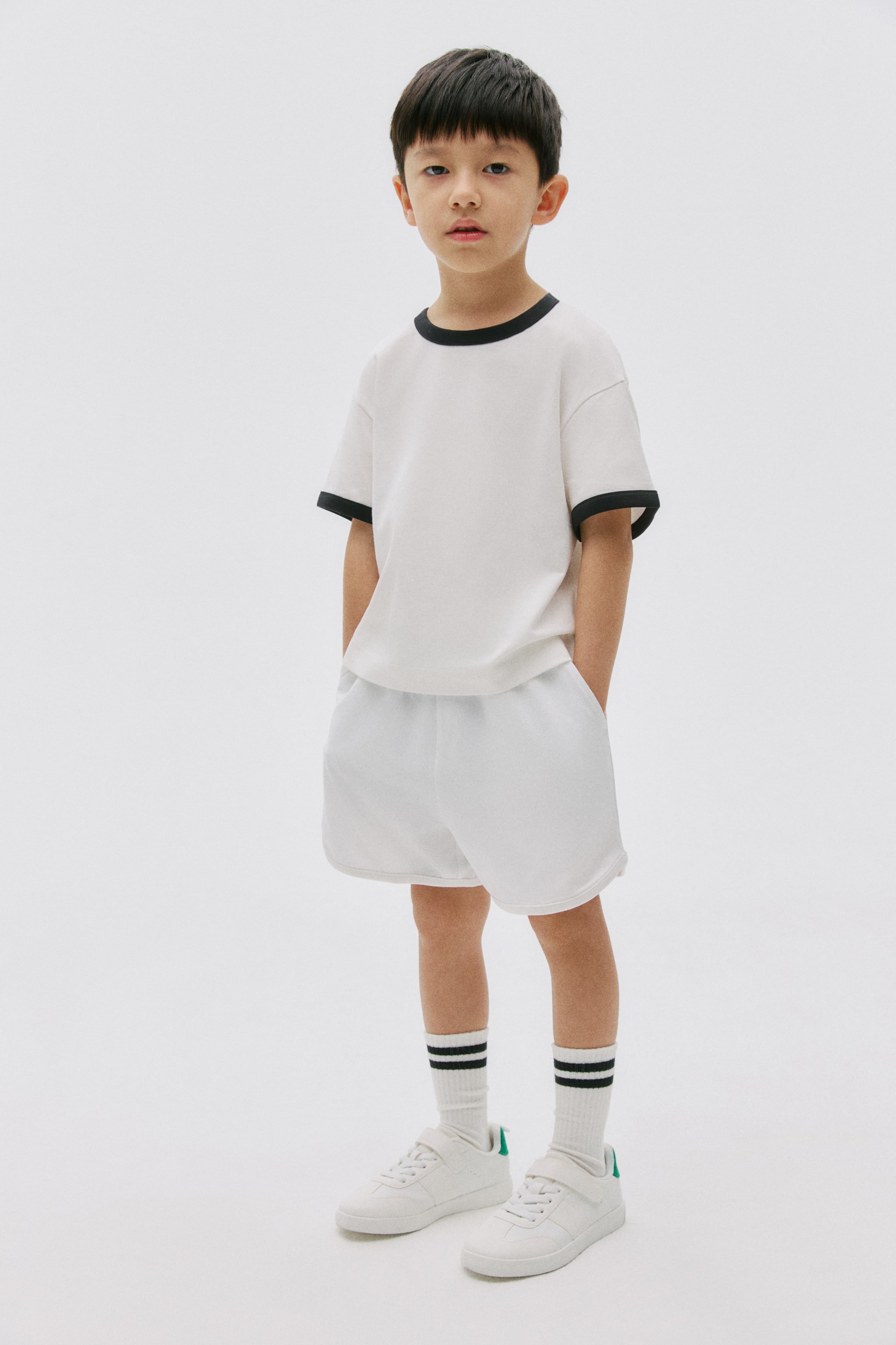 View larger image: A white short-sleeved t-shirt with a black crew neck and black sleeve trim is being worn by the model, paired with white shorts. The model's hands are resting in the shorts' pockets, while white striped socks and white sneakers are also being worn.