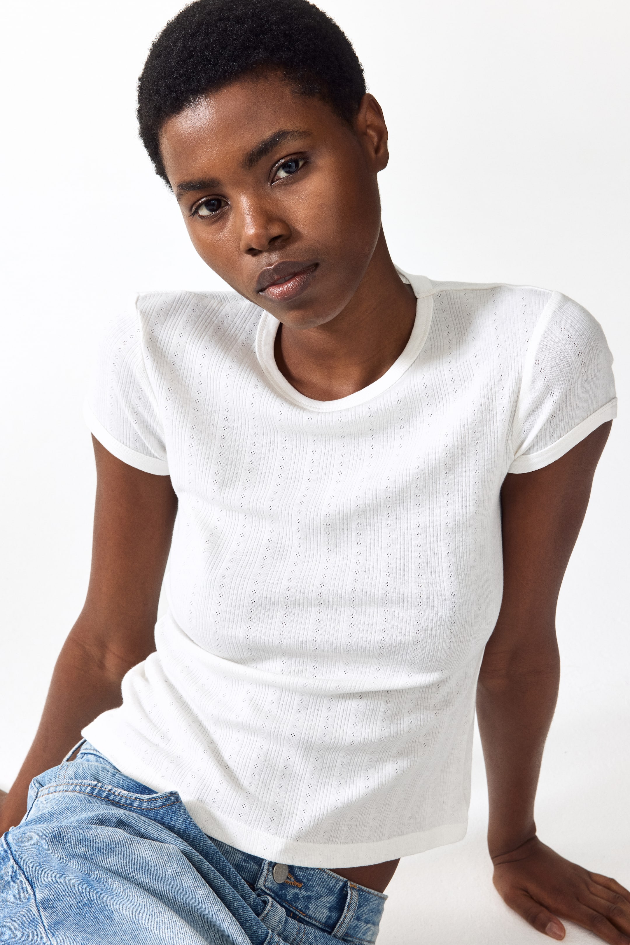 Se større billede: En tætsiddende t-shirt i fin pointelle-strikket bomuld med ribkant omkring halsudskæringen og ved manchetterne bæres af modellen. T-shirten er stylet med blå denimjeans.